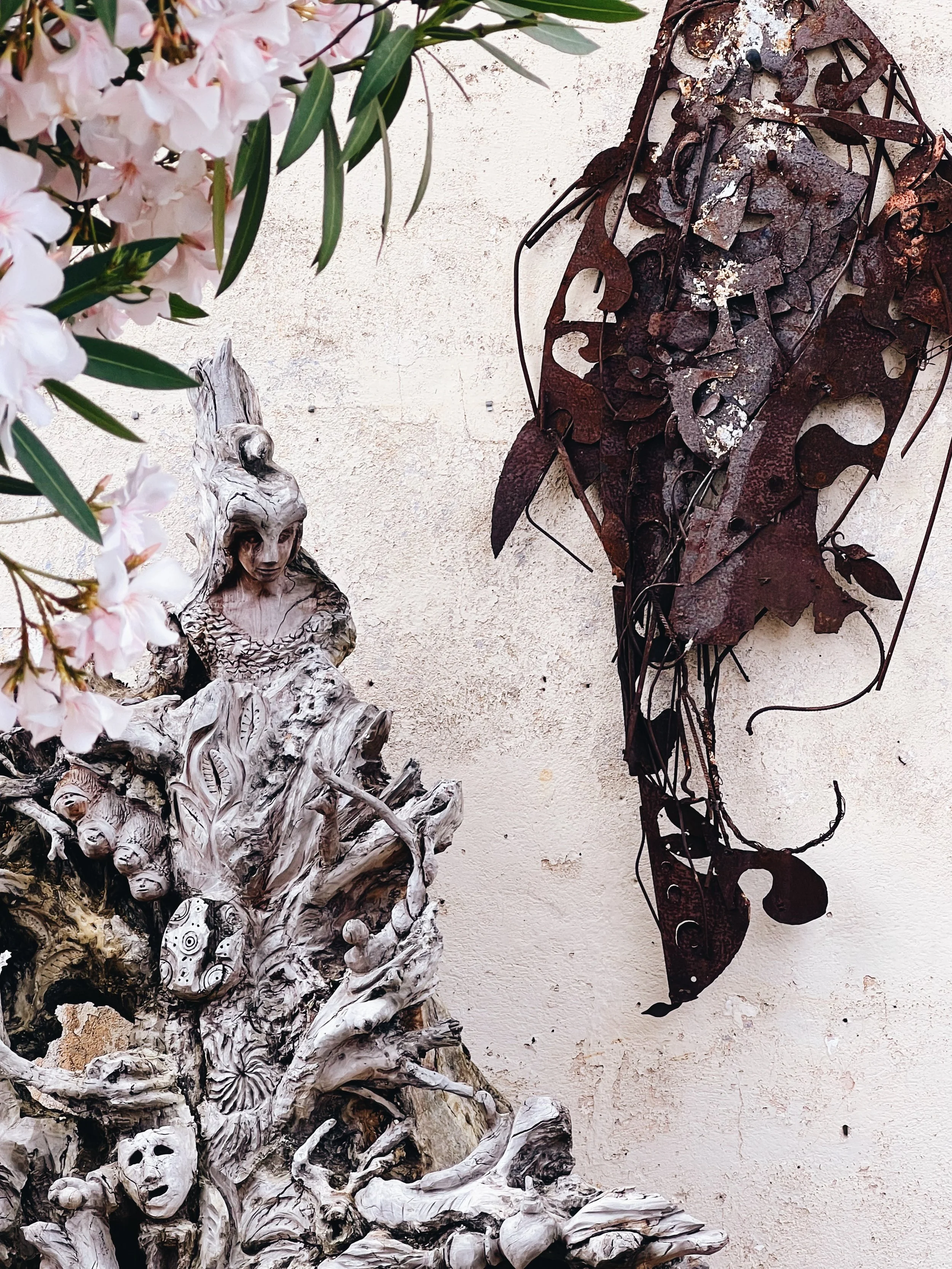 A wall-mounted art piece featuring a humanoid figure with a skeletal face and intricate details, surrounded by a textured, bark-like structure, and a rusty metal abstract face sculpture to the right, with pink flowers and green leaves on the left sid