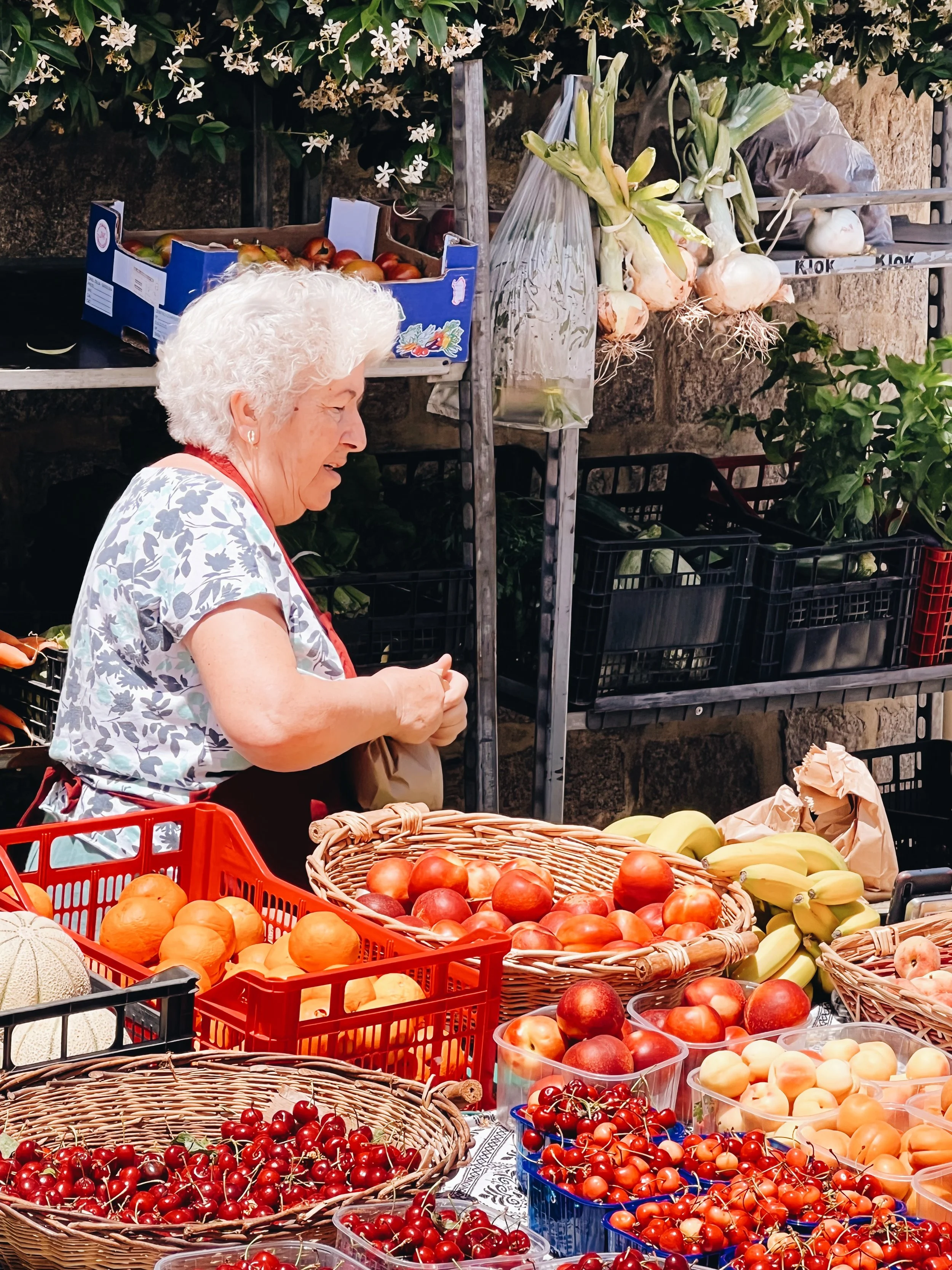 An elderly woman with curly white hair shopping at an outdoor farmer's market, looking at baskets of strawberries, peaches, bananas, and cherries on a table.