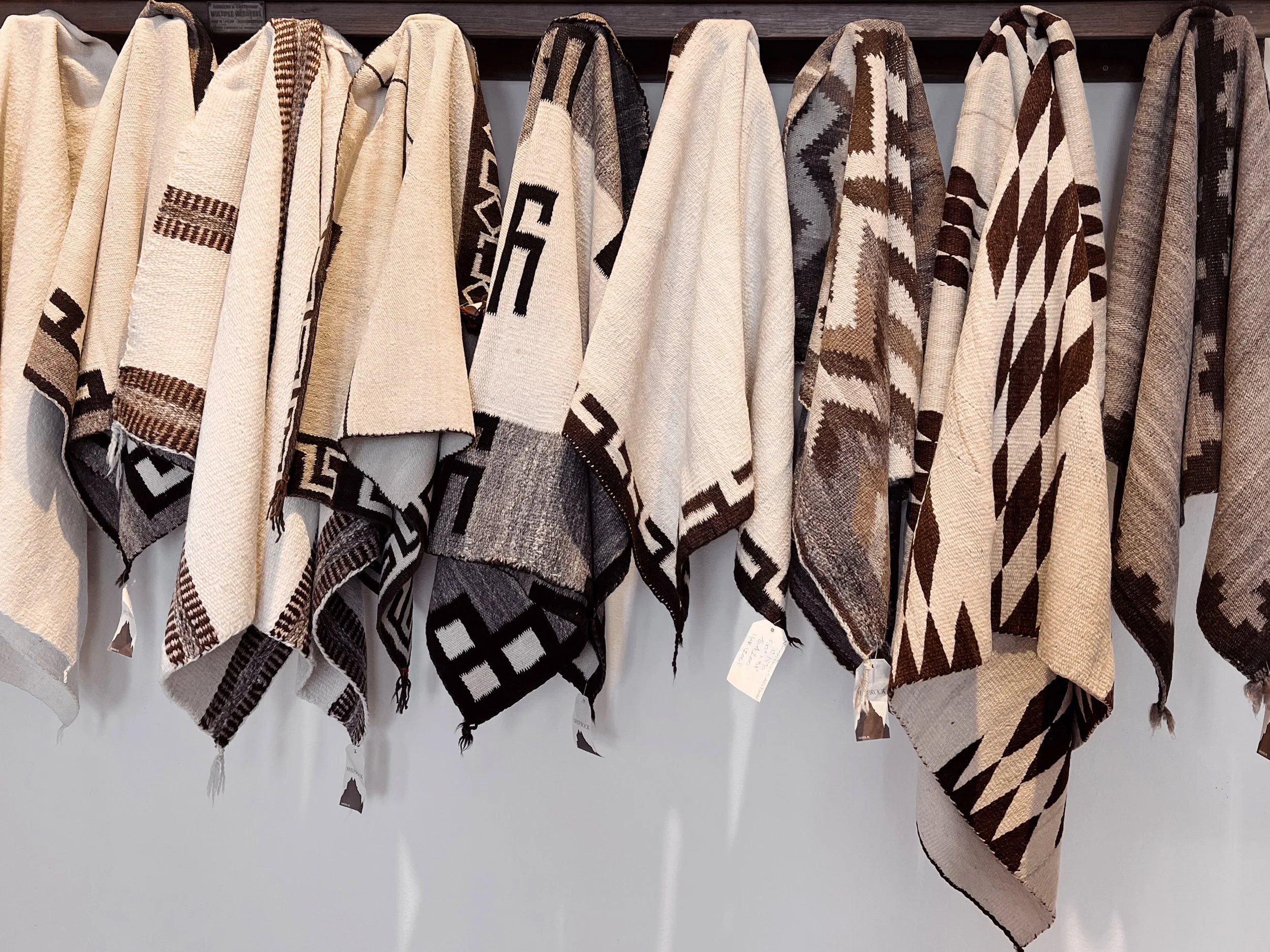 A row of shawls or blankets with geometric patterns in shades of beige, brown, black, and white, hanging on a rack against a wood-paneled background.