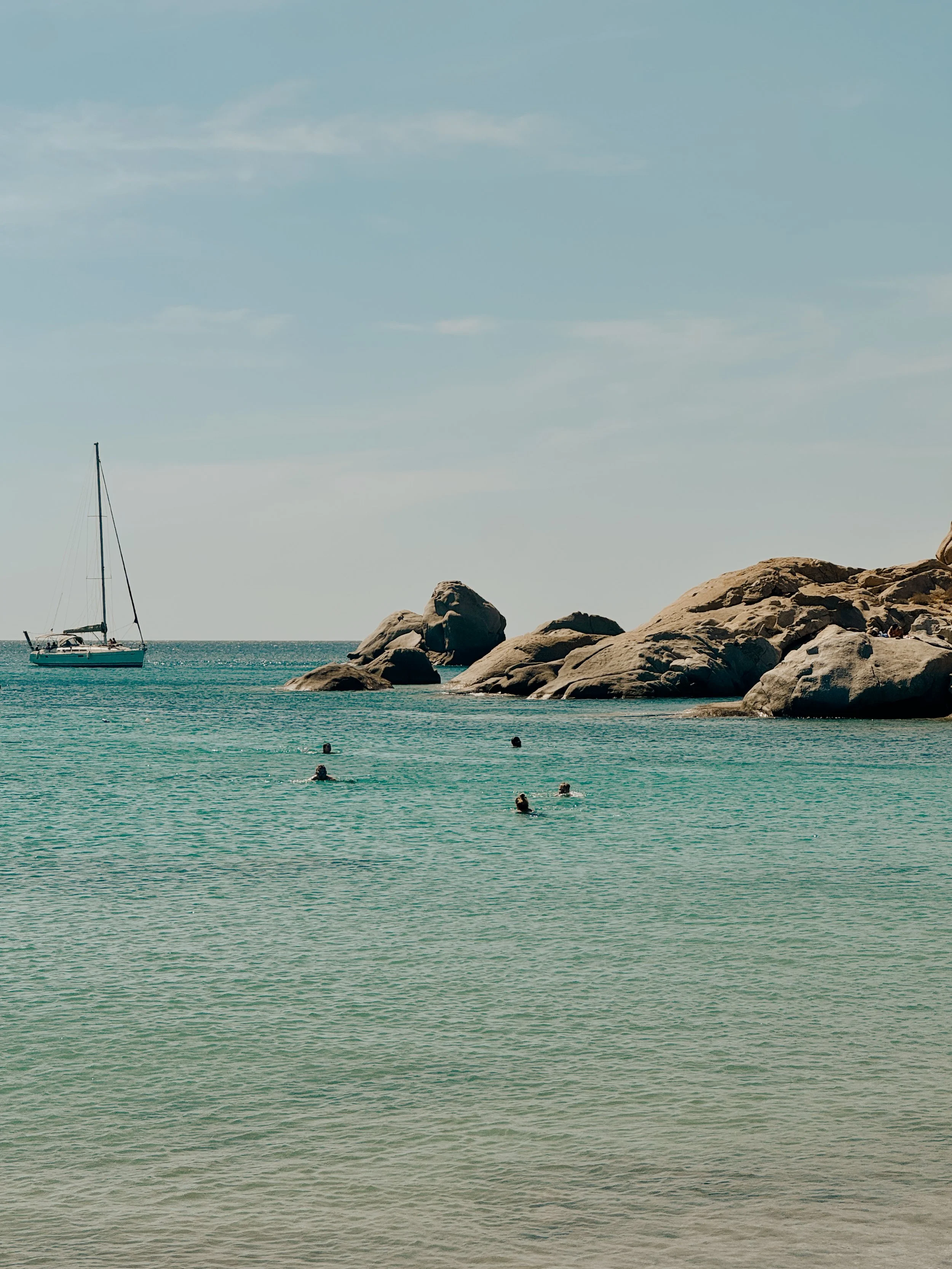 A calm ocean with a sailboat near rocks and several swimmers in the water under a partly cloudy sky.