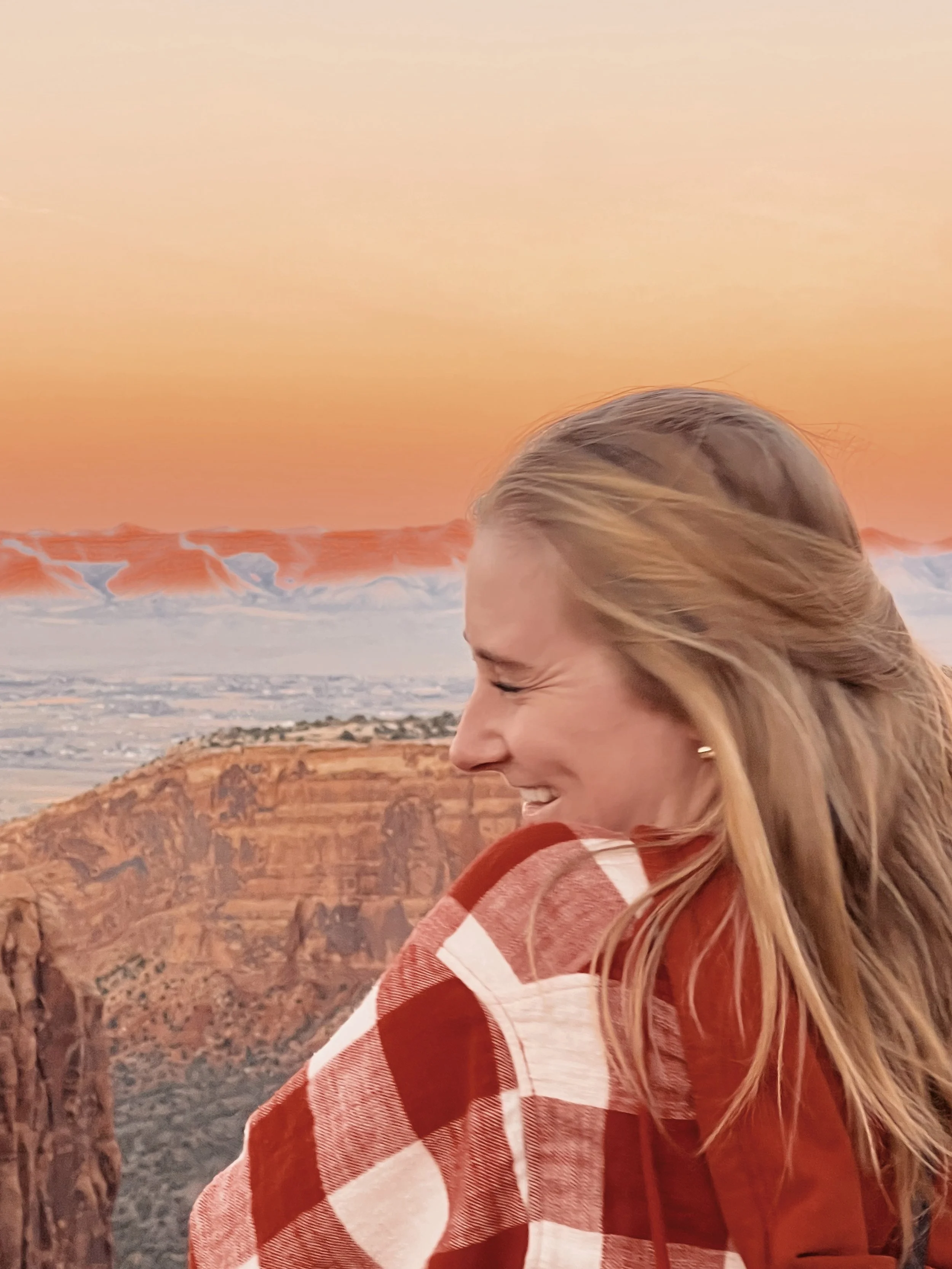A woman with blonde hair smiling against a sunset sky with mountains and a canyon in the background.