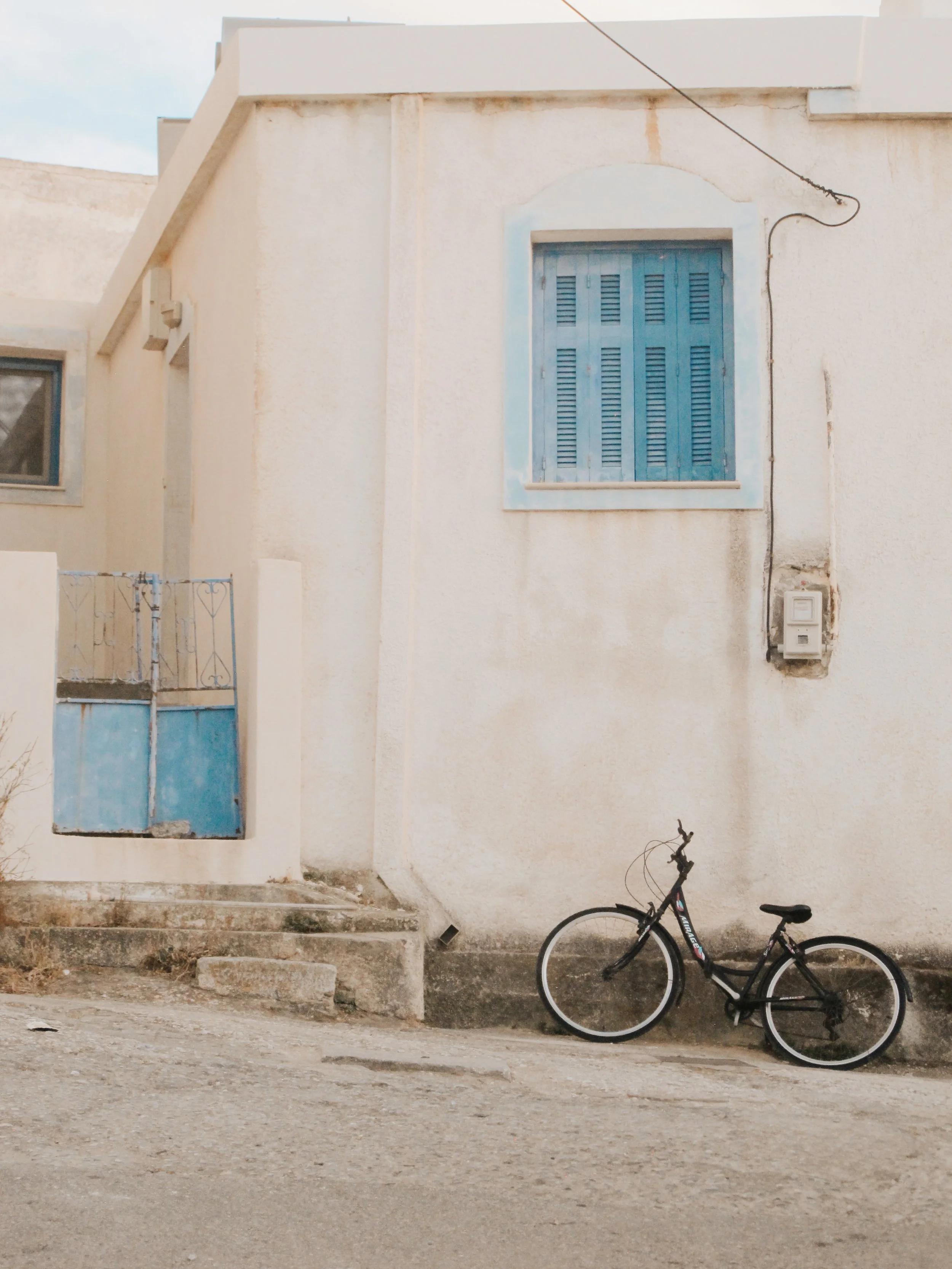 A black bicycle parked on a dirt ground in front of a beige building with two windows, one with blue shutters, and a small blue door with a decorative metal railing.