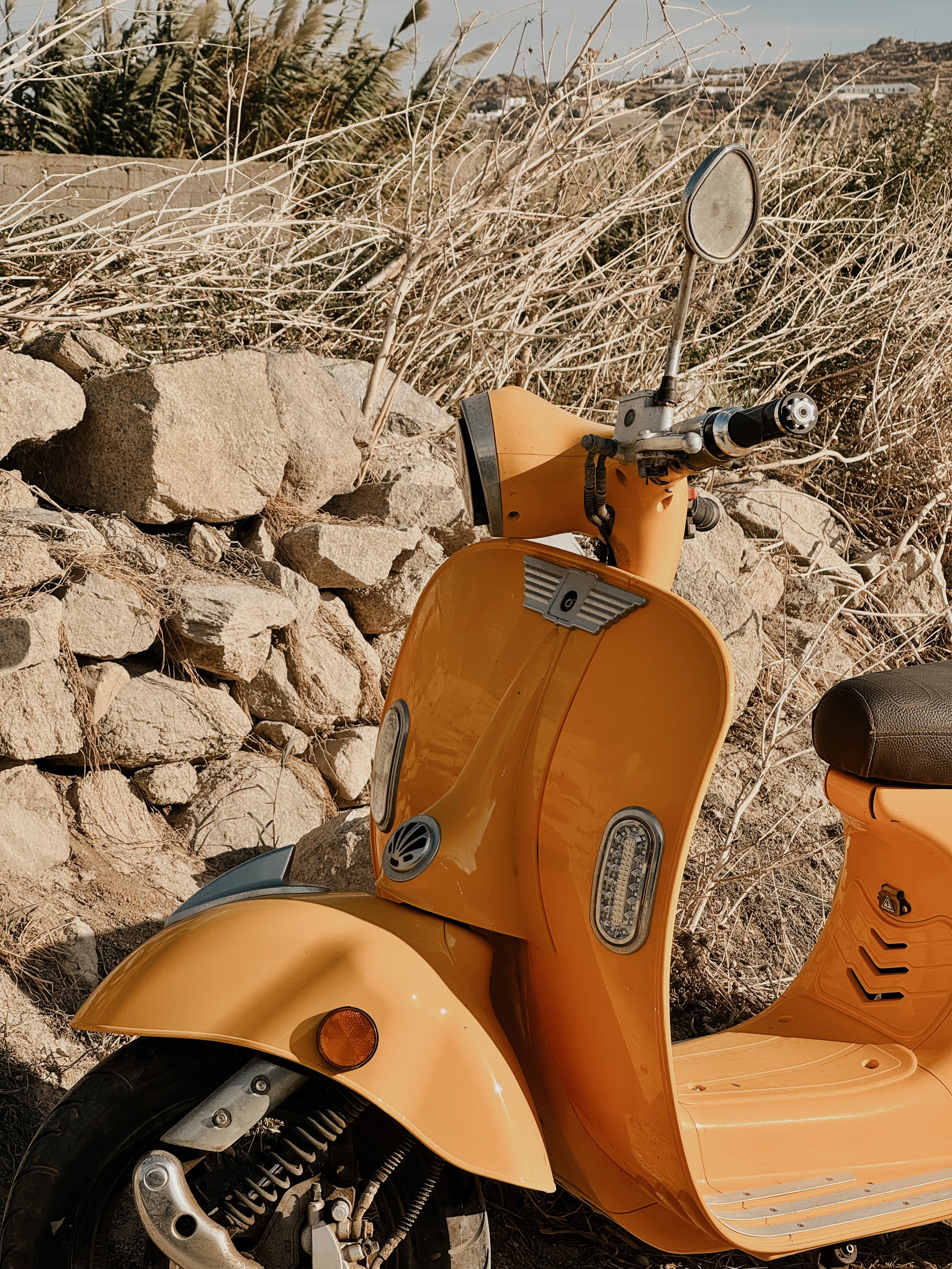 A bright orange scooter parked against a rocky, dry landscape with sparse vegetation and a distant hillside with some buildings.