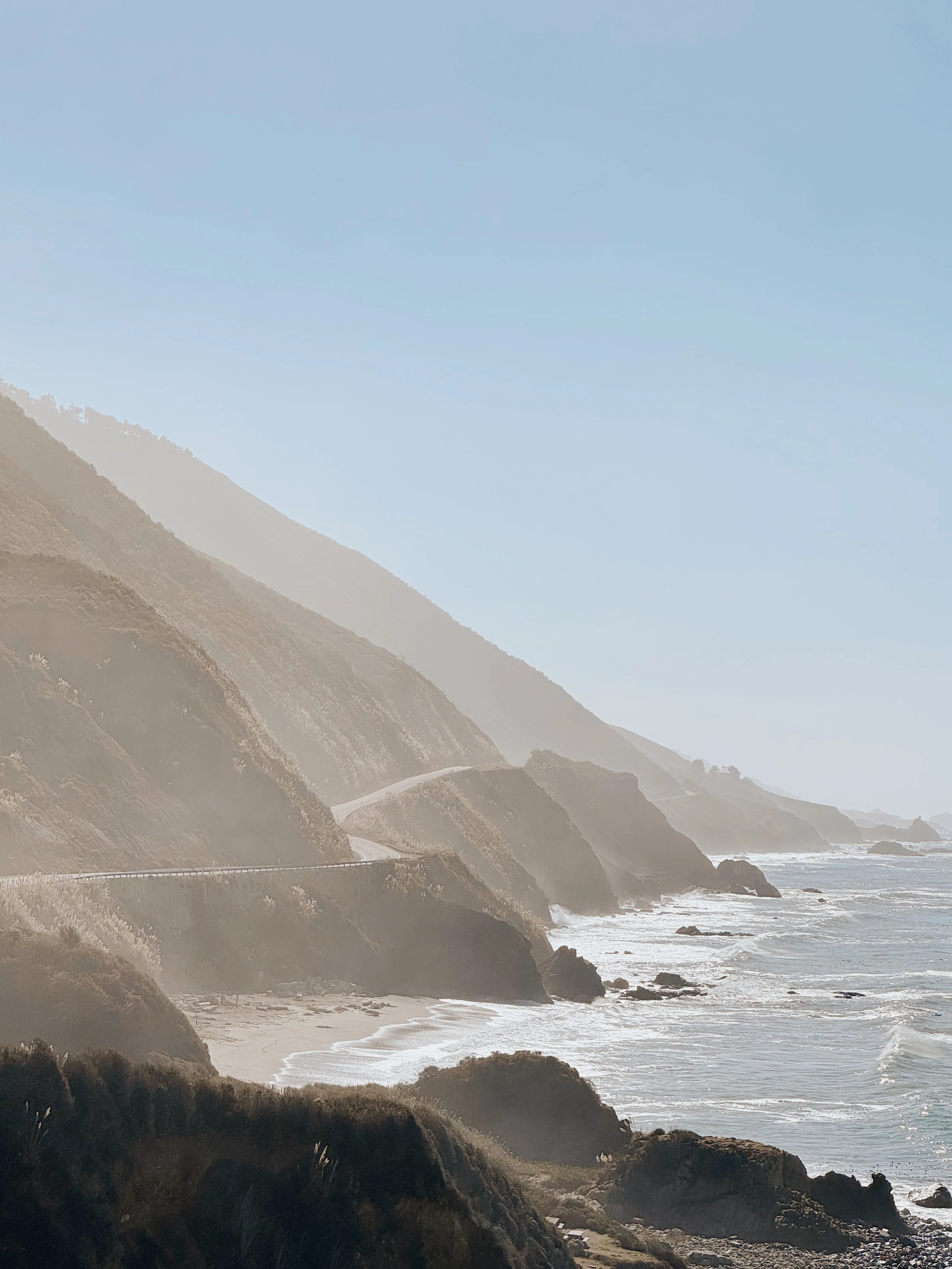 A scenic coastal view of rugged cliffs along a beach, with waves crashing against rocks and a winding road cutting through the hillside under a blue sky.