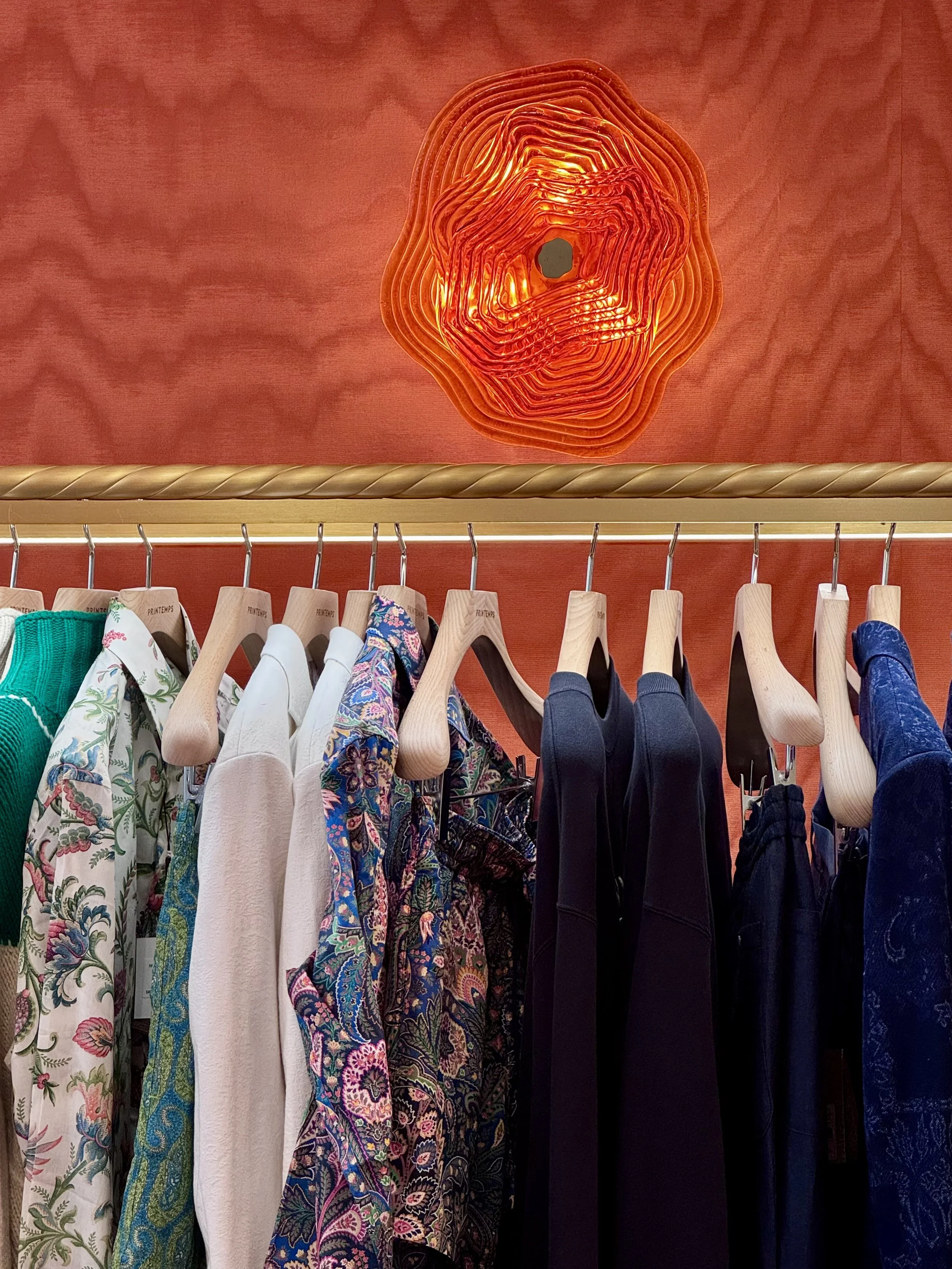 Clothing items hanging on a rack in a store, with a decorative orange light fixture on a reddish-orange wall above.