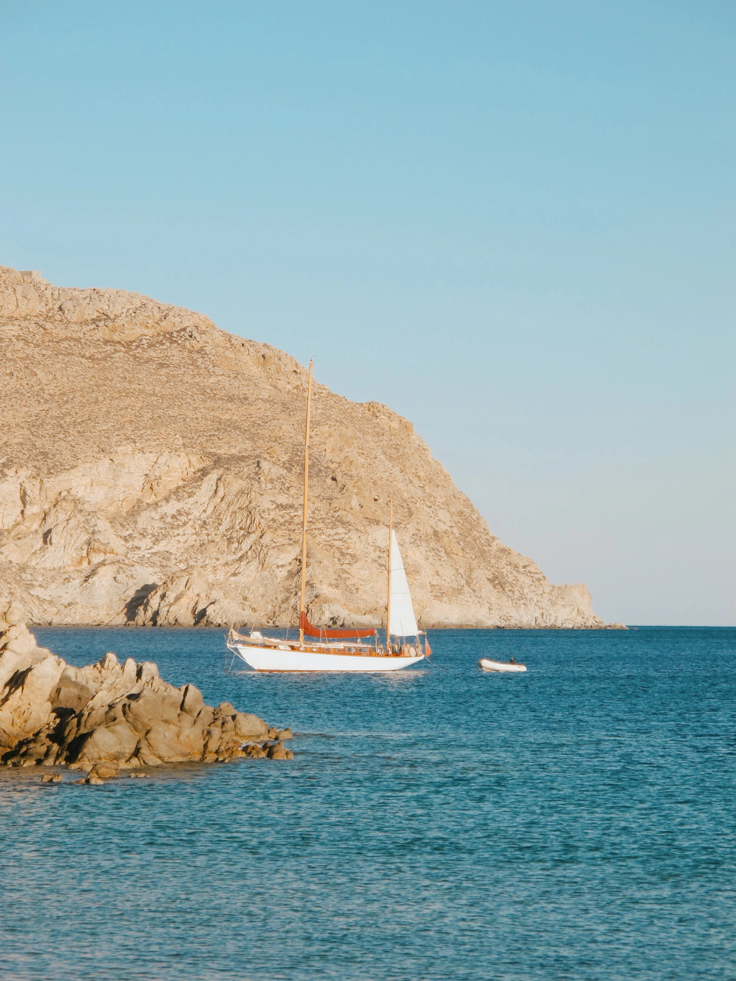 A sailboat with white sails floating in blue water near rocky coastline with a large, rugged mountain in the background under a clear sky.