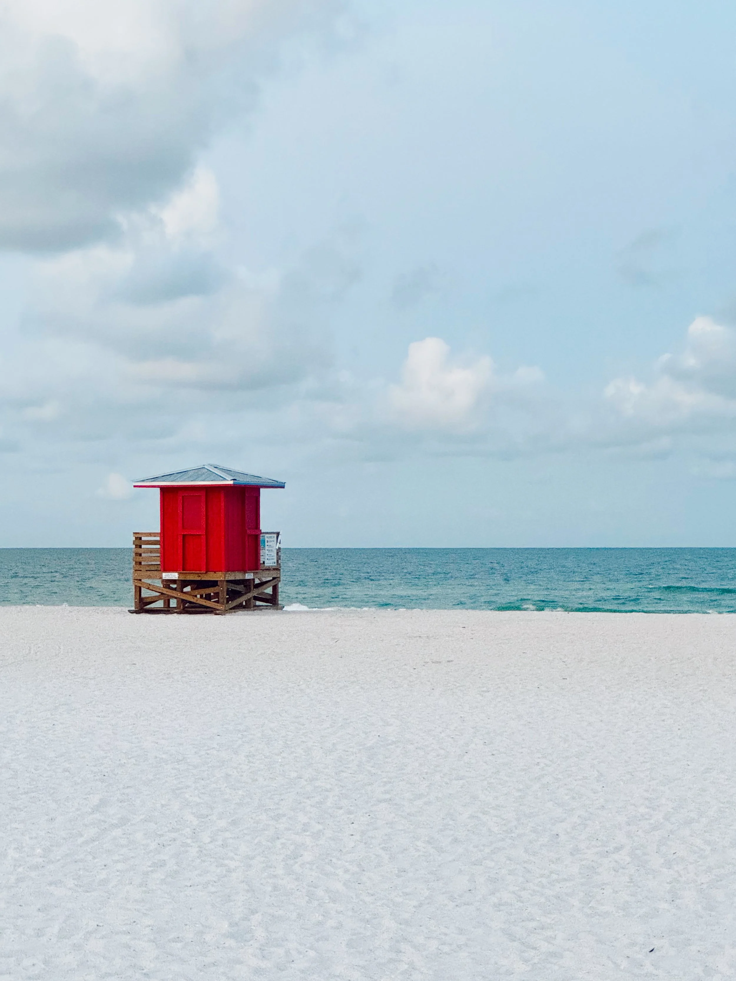 A red lifeguard station on a white sandy beach with the ocean and cloudy sky in the background.