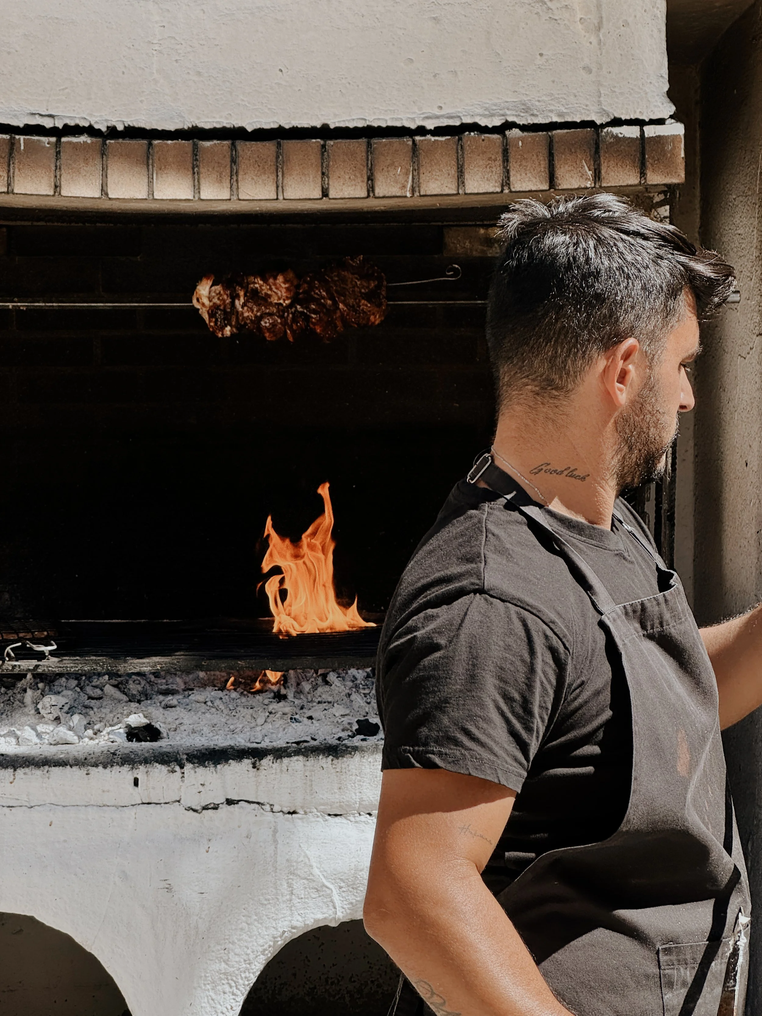 A man sitting in front of a barbecue grill with a steak on the fire. The man has a tattoo on his neck that reads 'Good vibes' and short, styled hair.