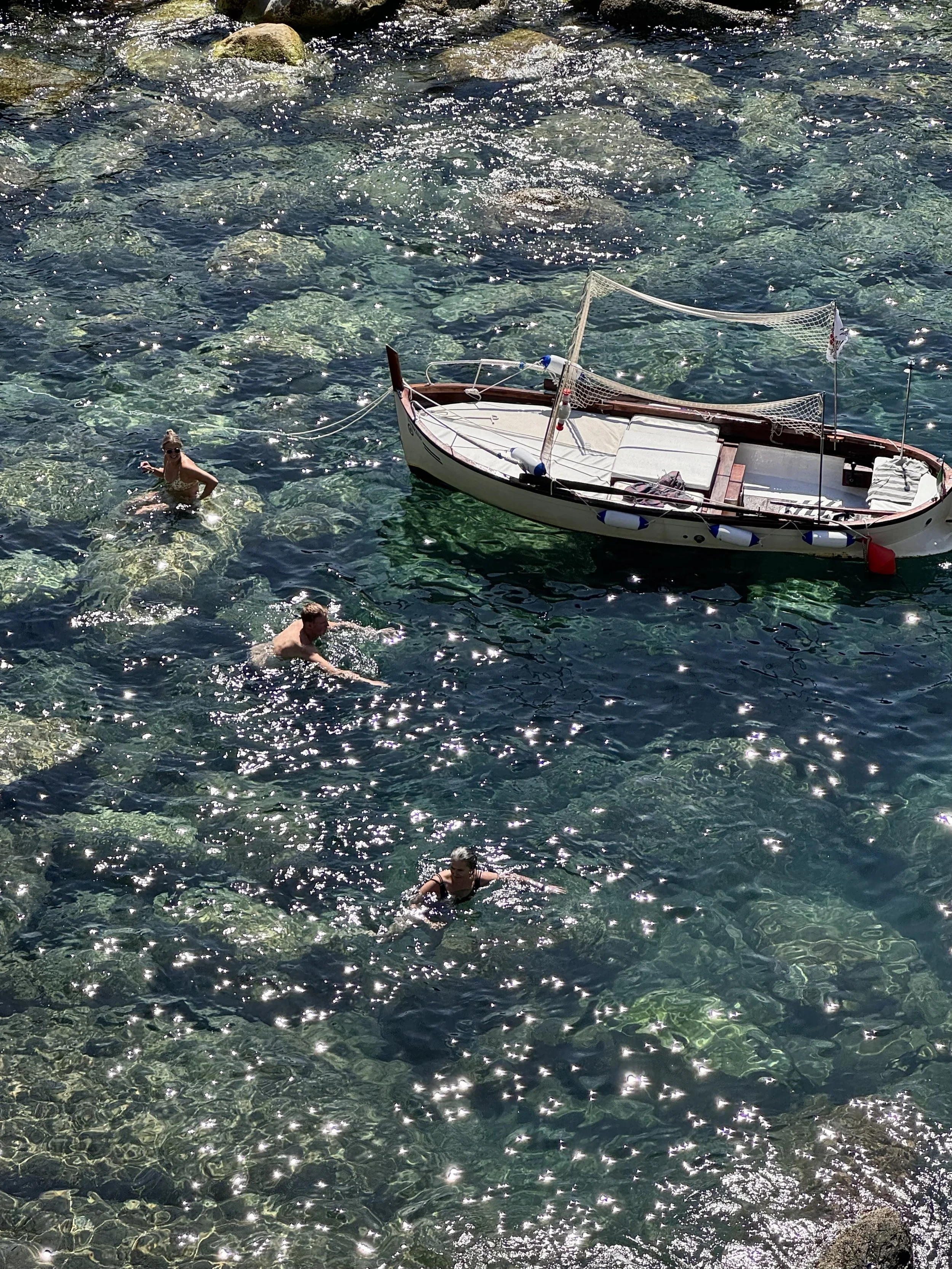 People swimming in clear water near a docked small boat with a net and striped cushion on the deck.