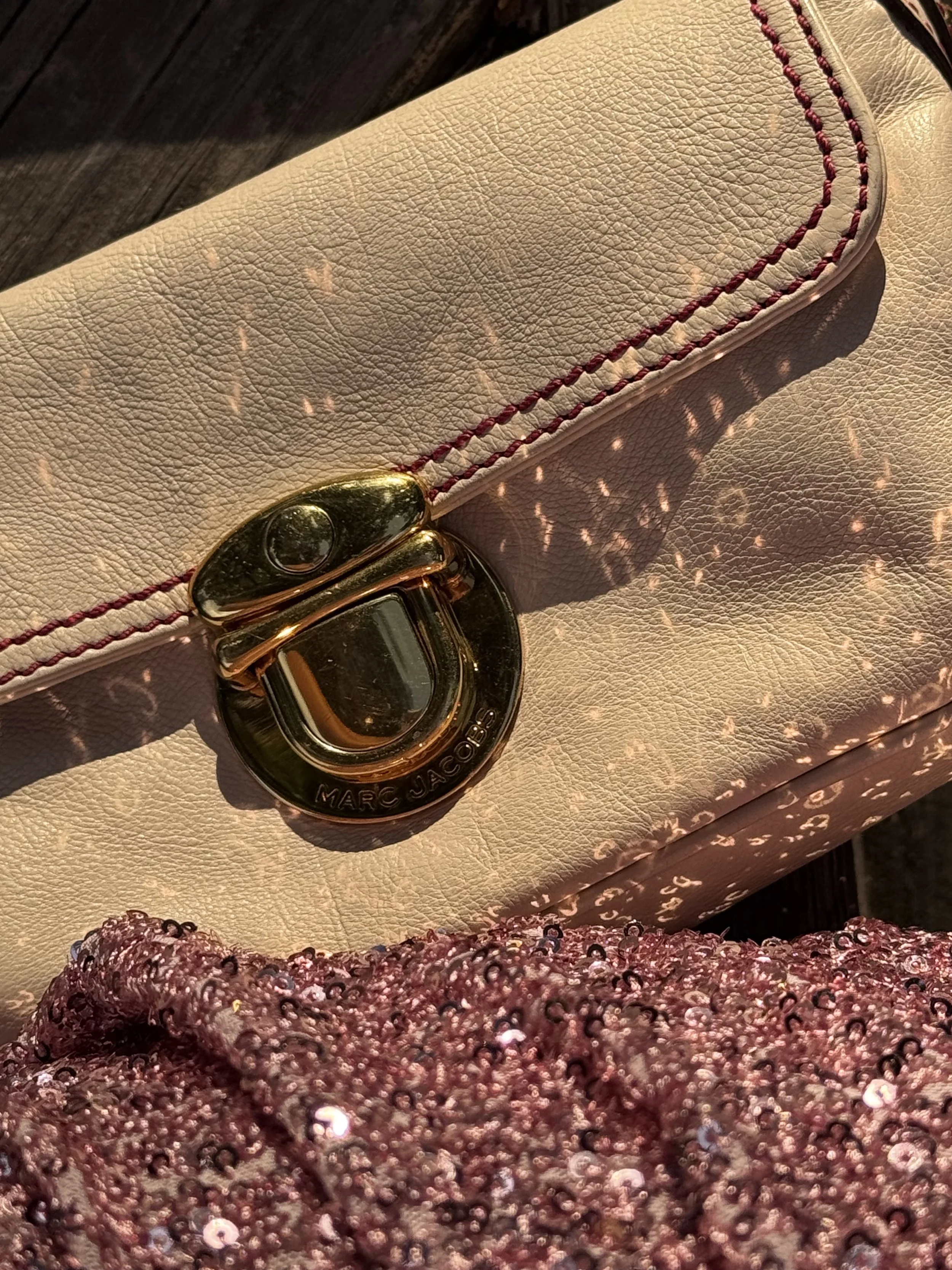Close-up of a cream-colored leather bag with red stitching and gold hardware, sunlight casting shimmering reflections on the surface.