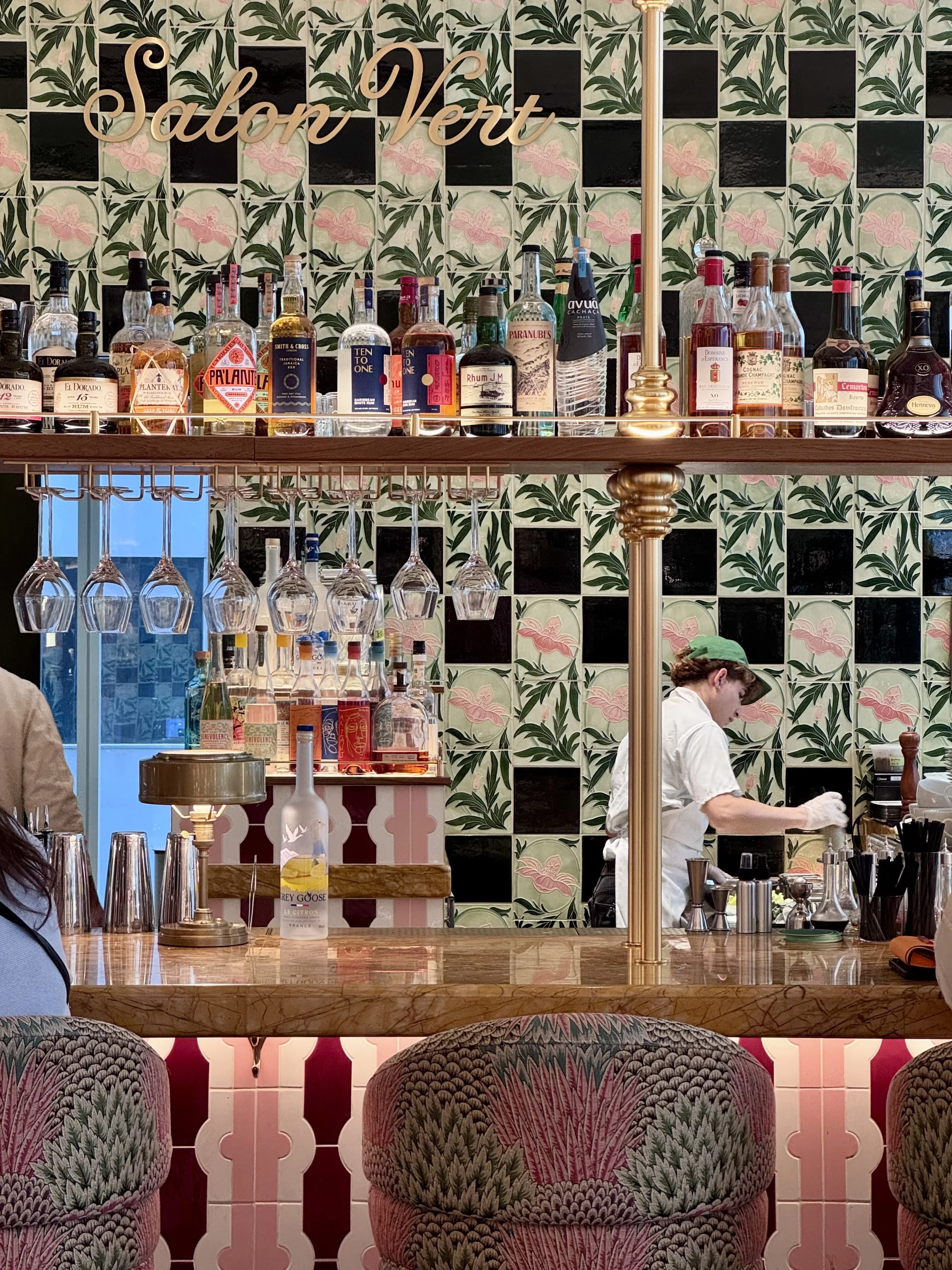 Bar scene at Salon Vert featuring a variety of liquor bottles on shelves, hanging wine glasses, and a bartender preparing drinks beneath a decorative floral wallpaper and a neon sign that says "Salon Vert."