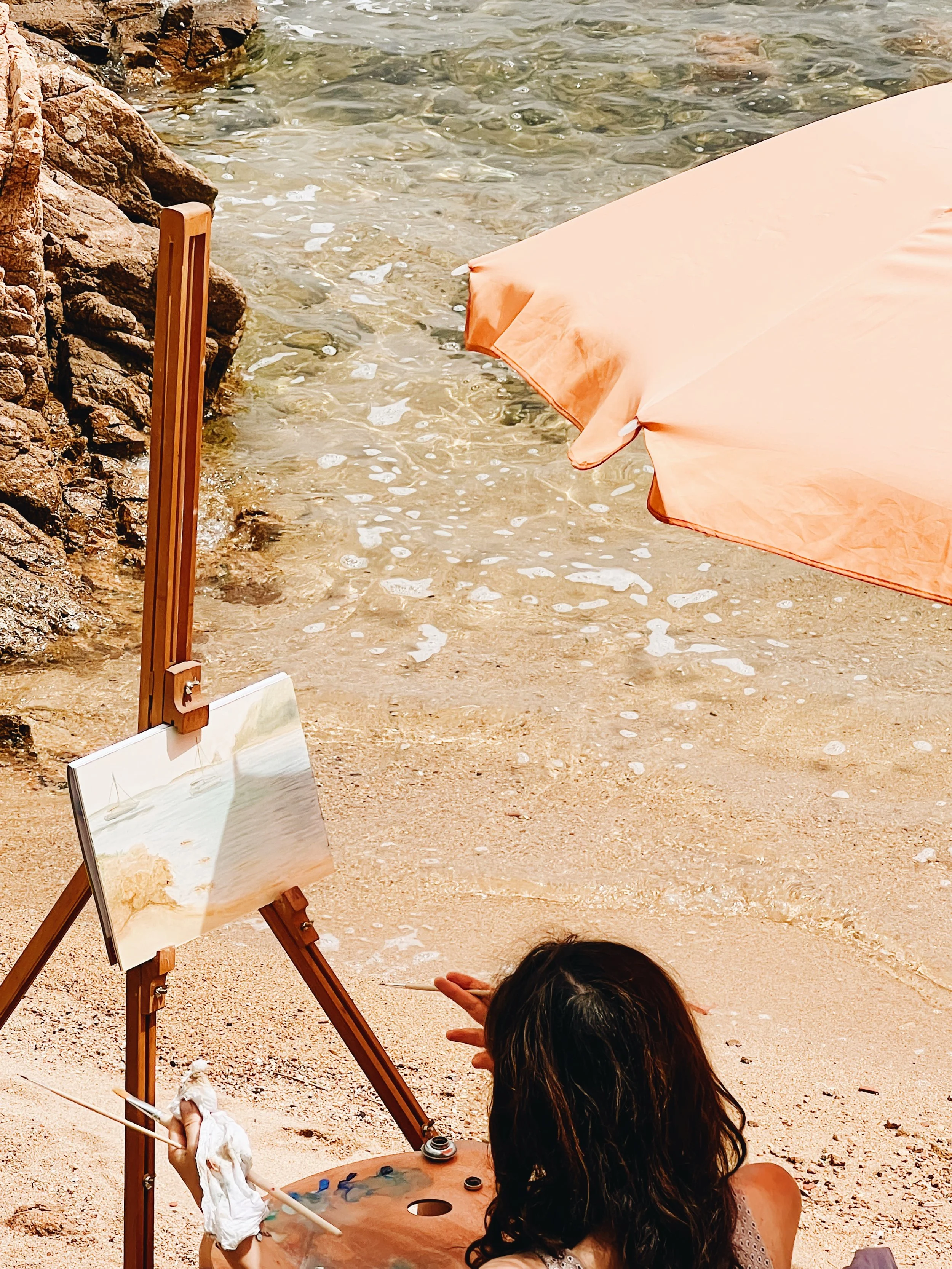 A person sitting on the beach with long dark hair, painting on a canvas placed on an easel, near the water's edge. The scene includes a beige umbrella, rocks, and clear shallow water with small waves.