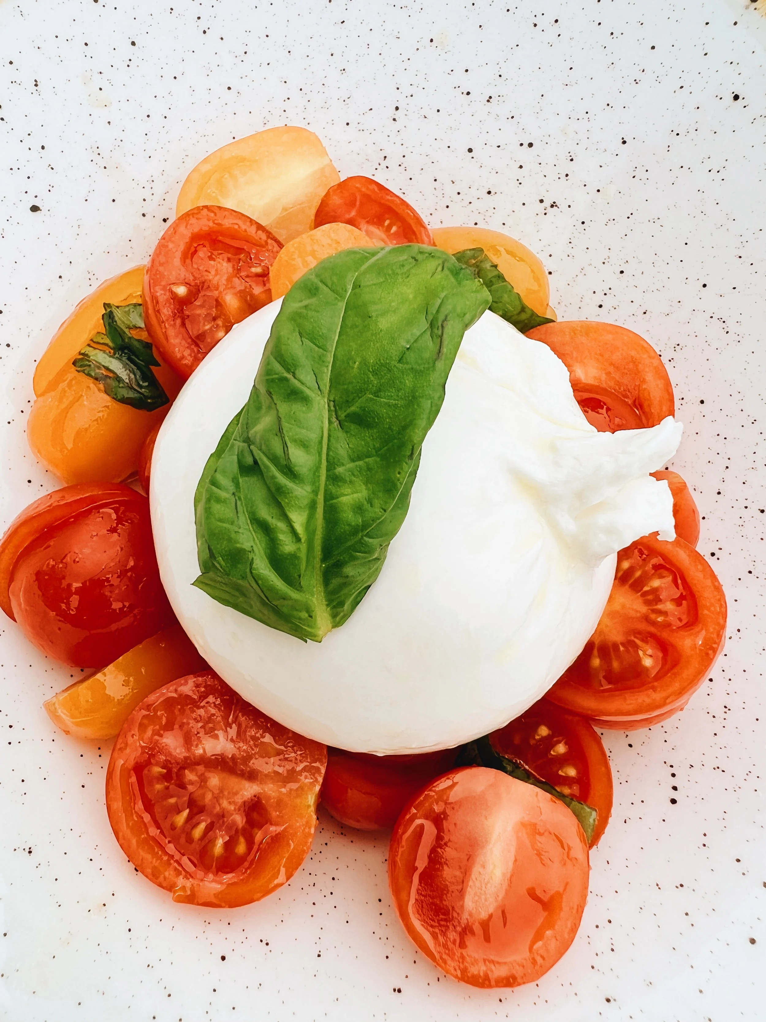 Sliced cherry tomatoes, a fresh basil leaf, and a ball of mozzarella cheese on a speckled white surface.
