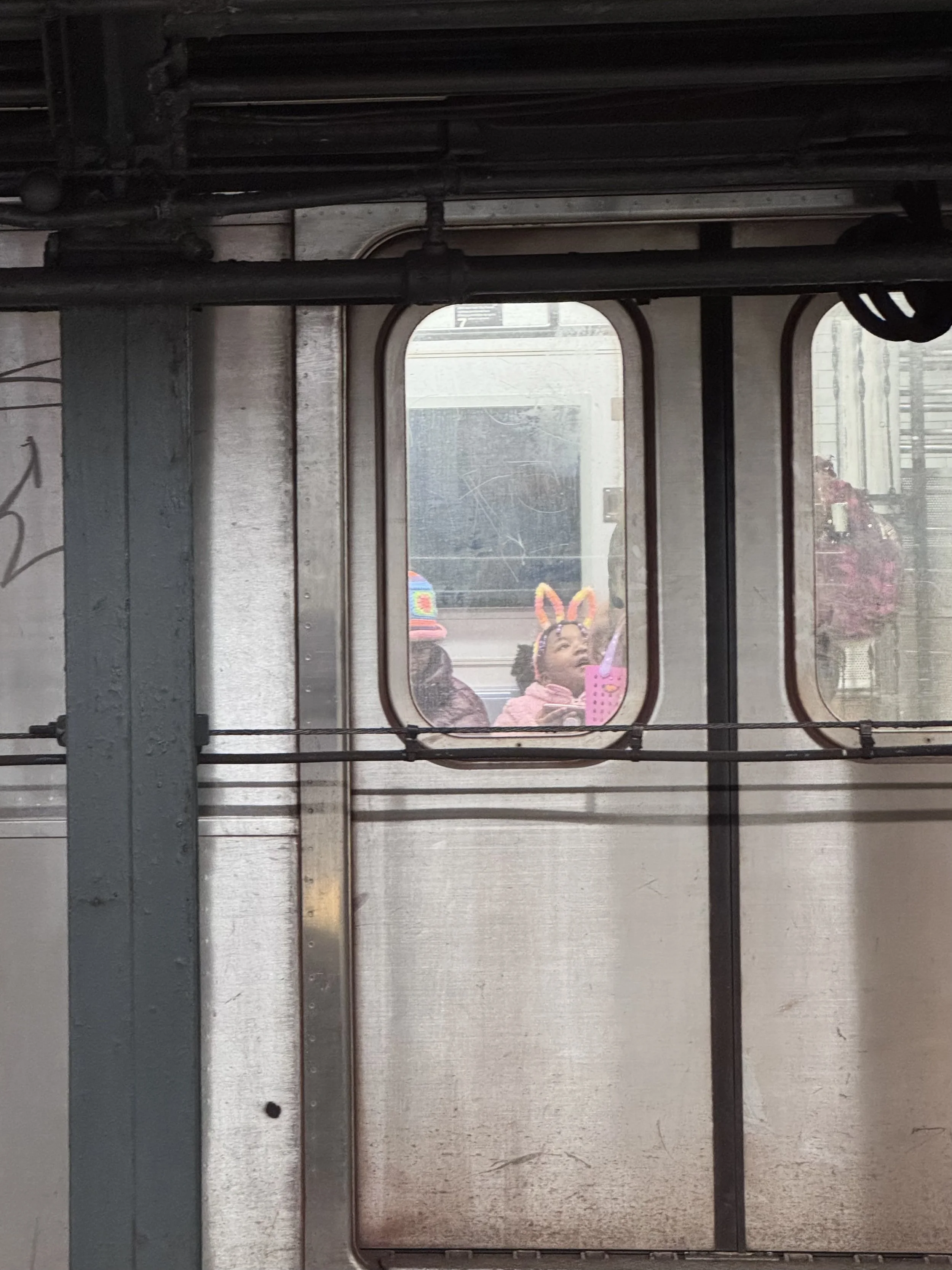 View through a window showing a woman with bunny ears and pink antennas, holding a pink item, with colorful balloons in the background.