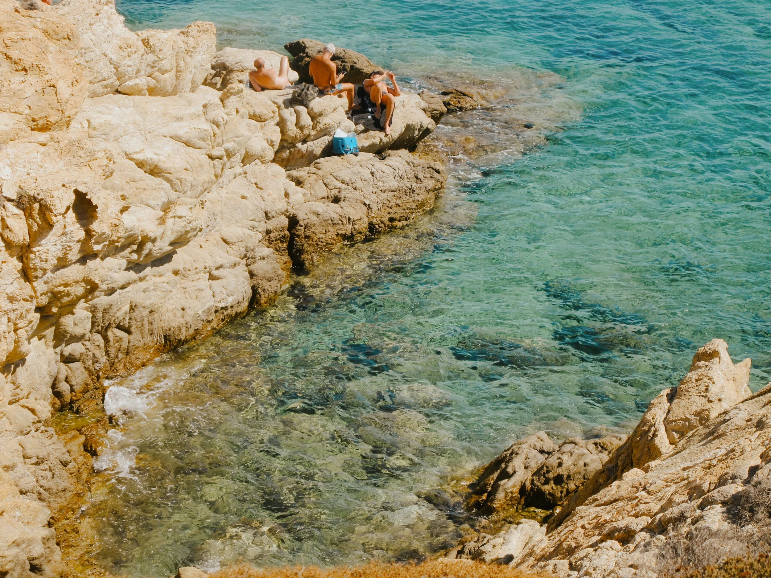 Three men sitting on rocks at the edge of the ocean, relaxing and one using a smartphone, with clear turquoise water and rocky shoreline.