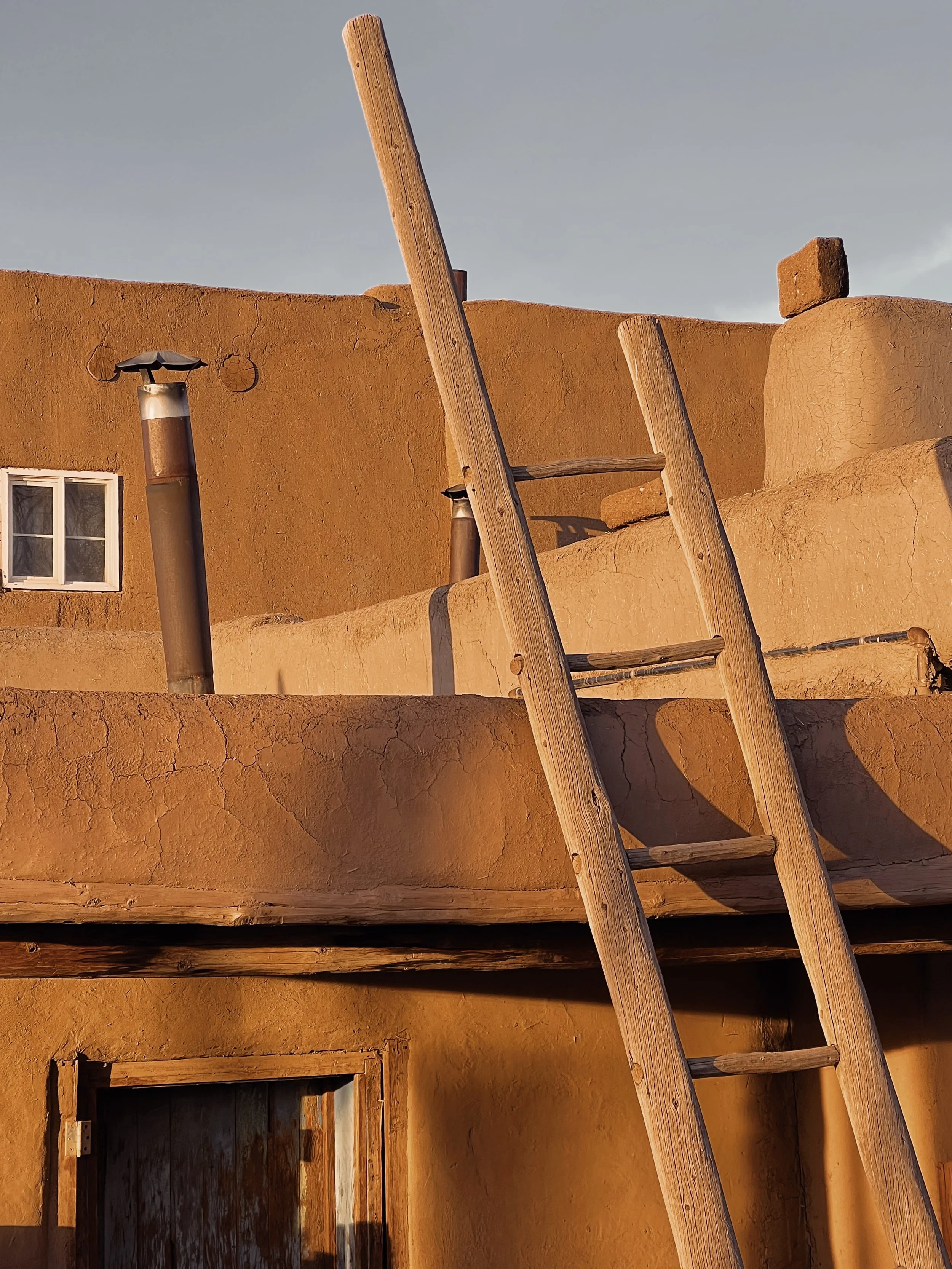 An adobe-style building with a ladder leaning against it, a small window, and two smoke vents on the roof under a clear sky.