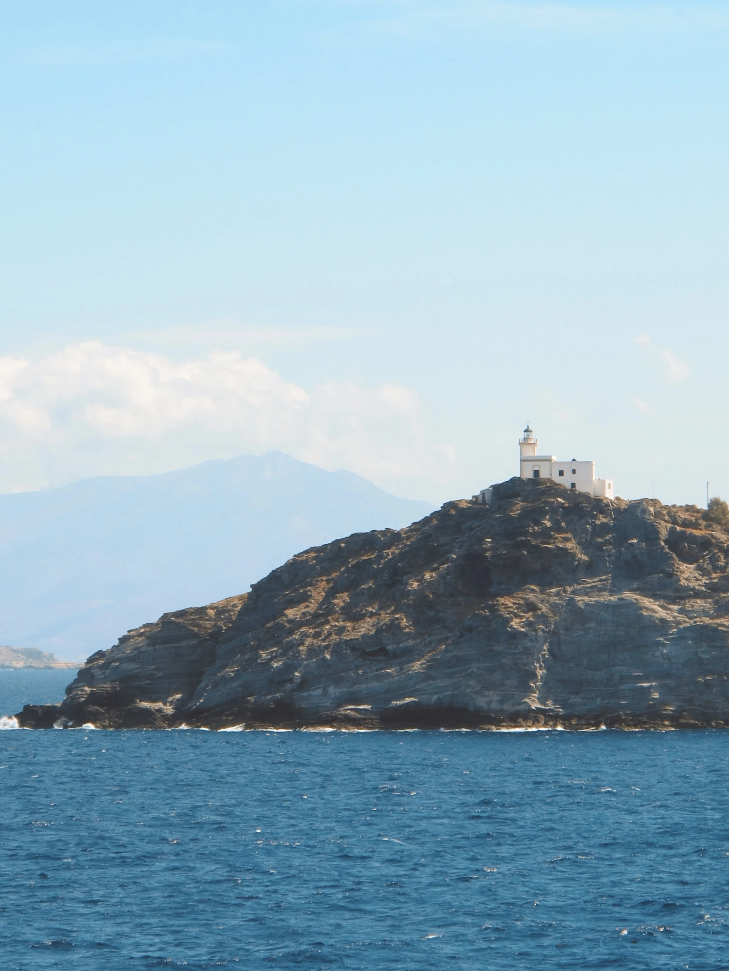 A lighthouse on a rocky hill overlooking the ocean with mountains in the background under a partly cloudy sky.