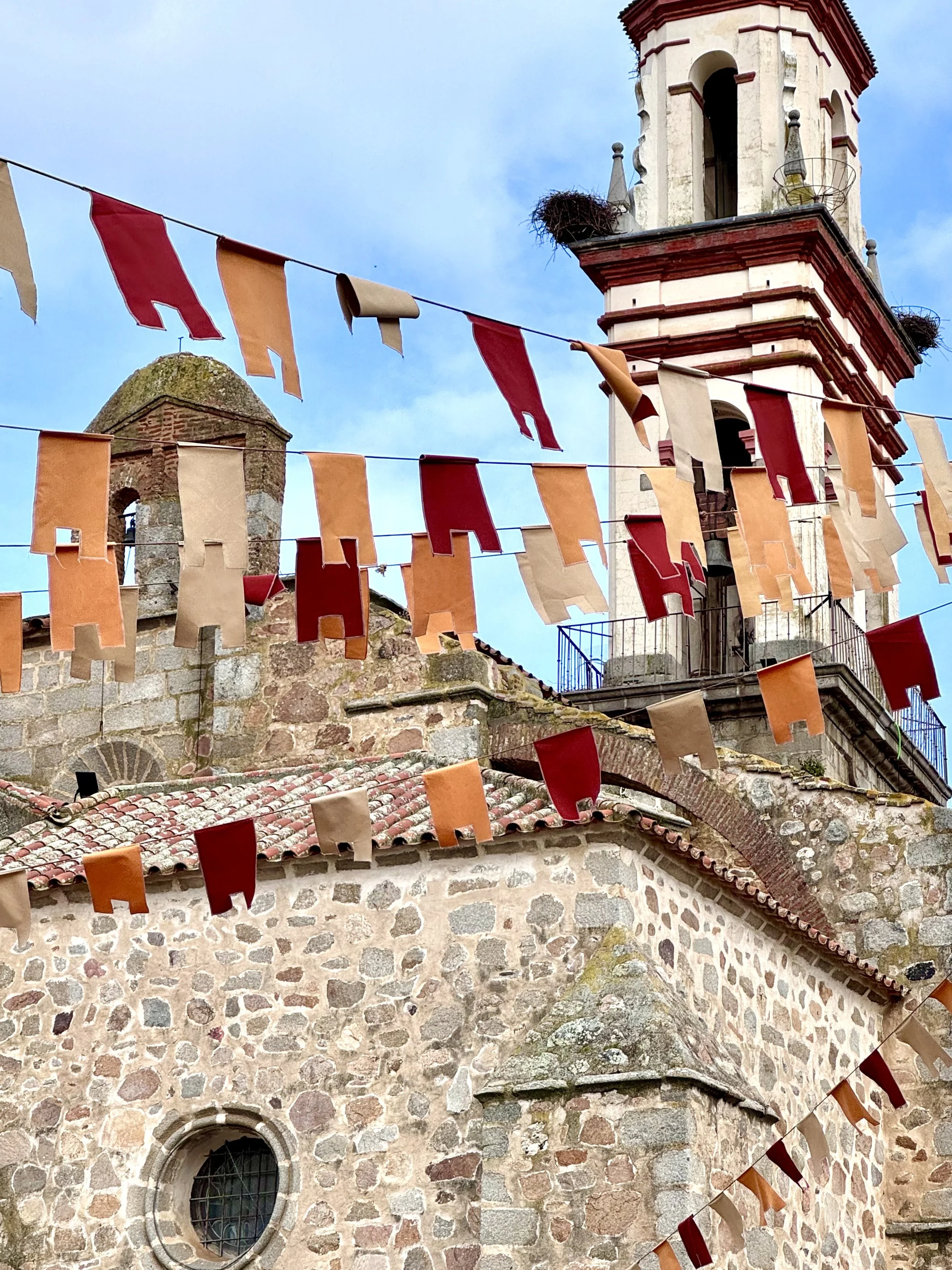 Colorful flags hanging across a stone building with a tower and a small circular window with bars, under a cloudy sky.