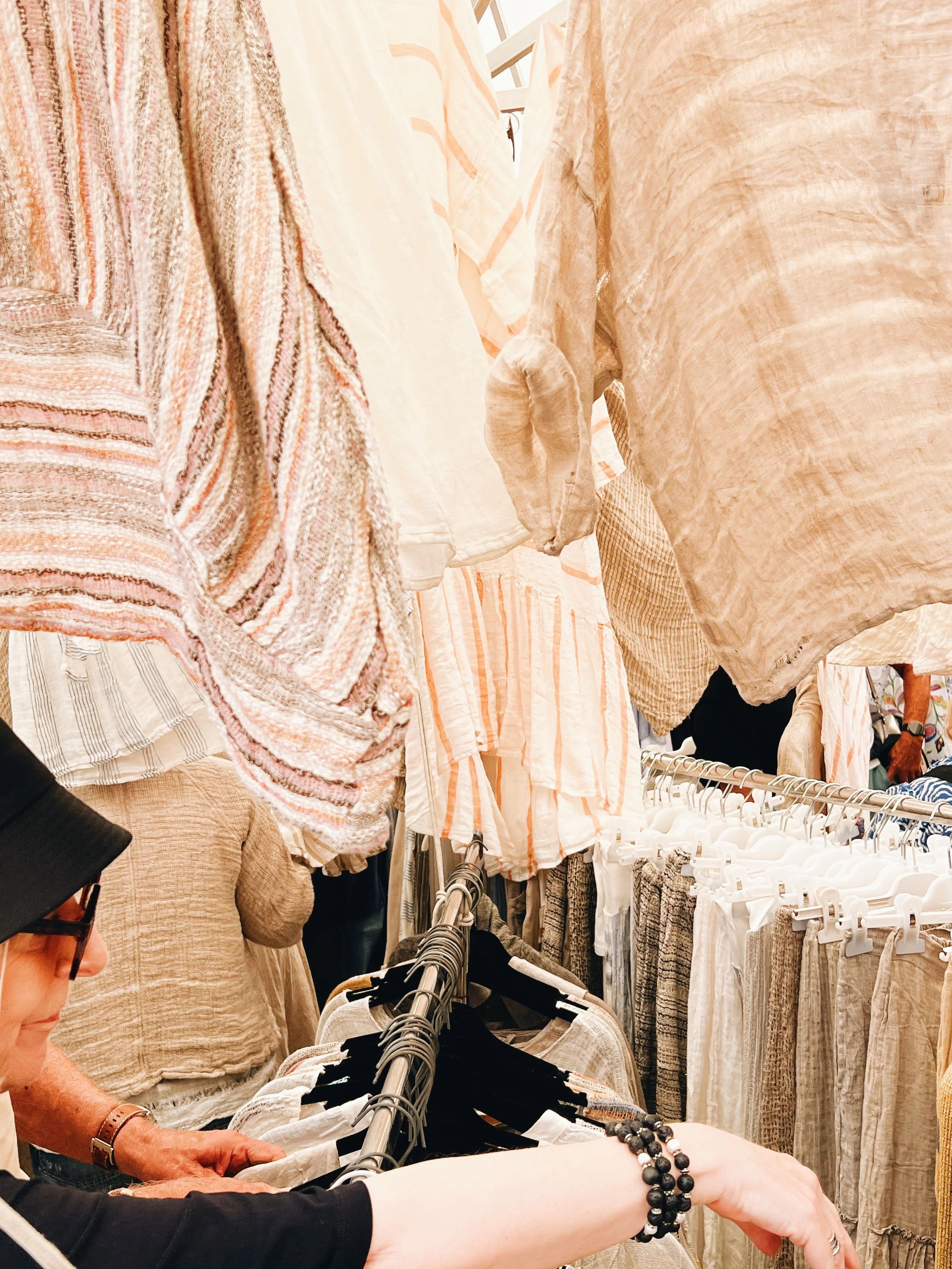 People shopping for clothing in a store, browsing racks with beige, peach, and cream-colored dresses, and woven fabrics in the background.