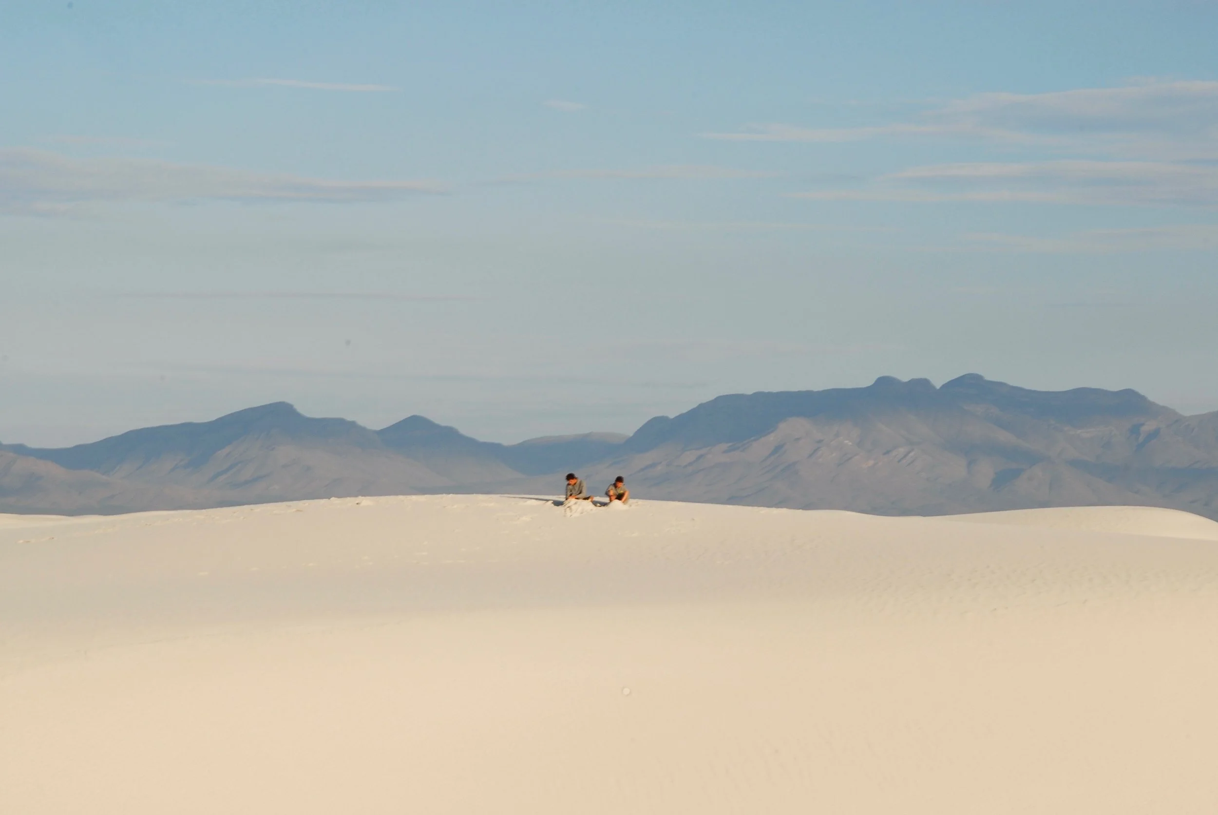 Two people sitting on a sandy landscape with mountains in the background under a blue sky.
