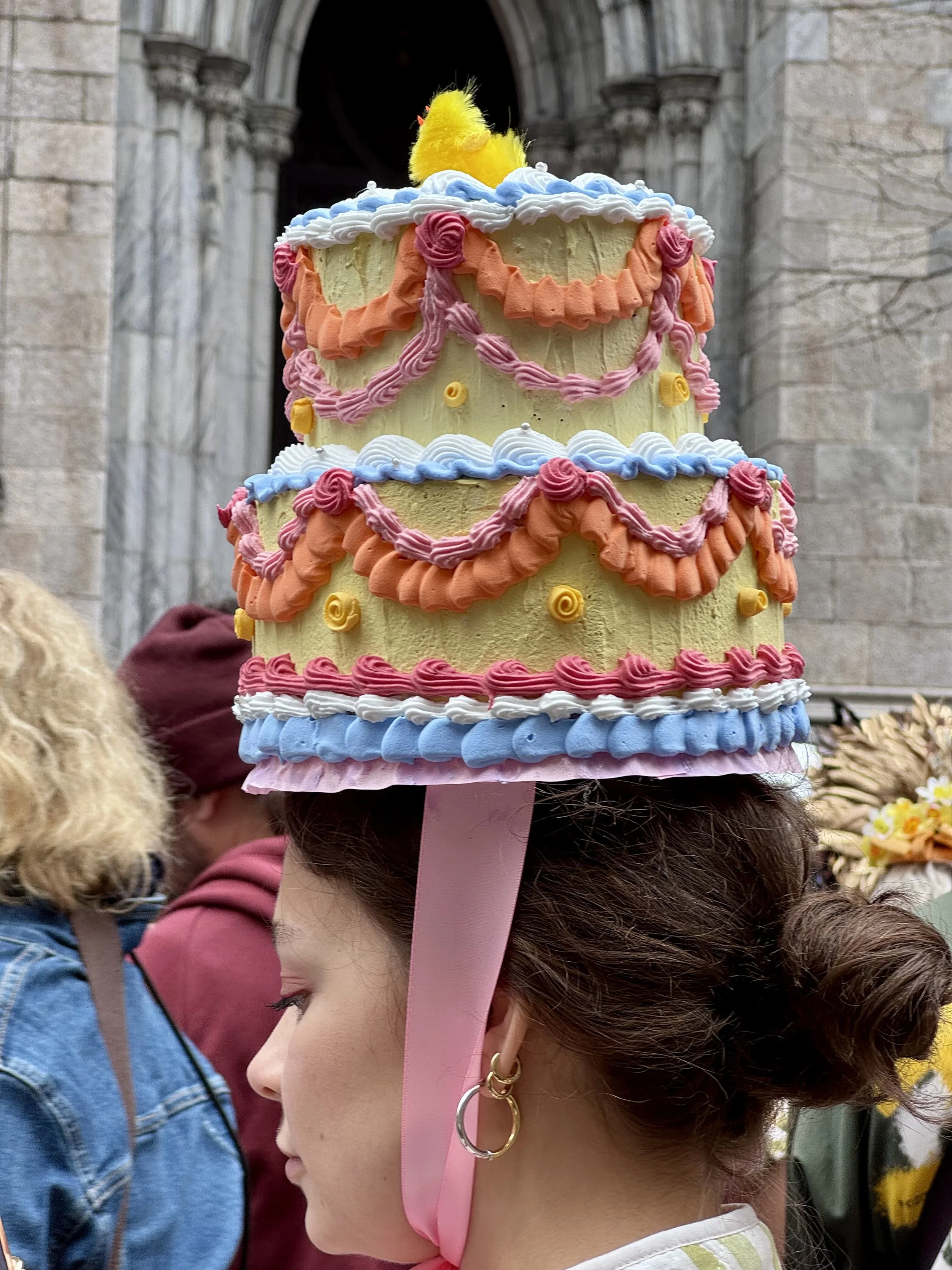 A woman wearing a decorative, tiered hat resembling a cake with colorful icing and a small yellow chick on top.