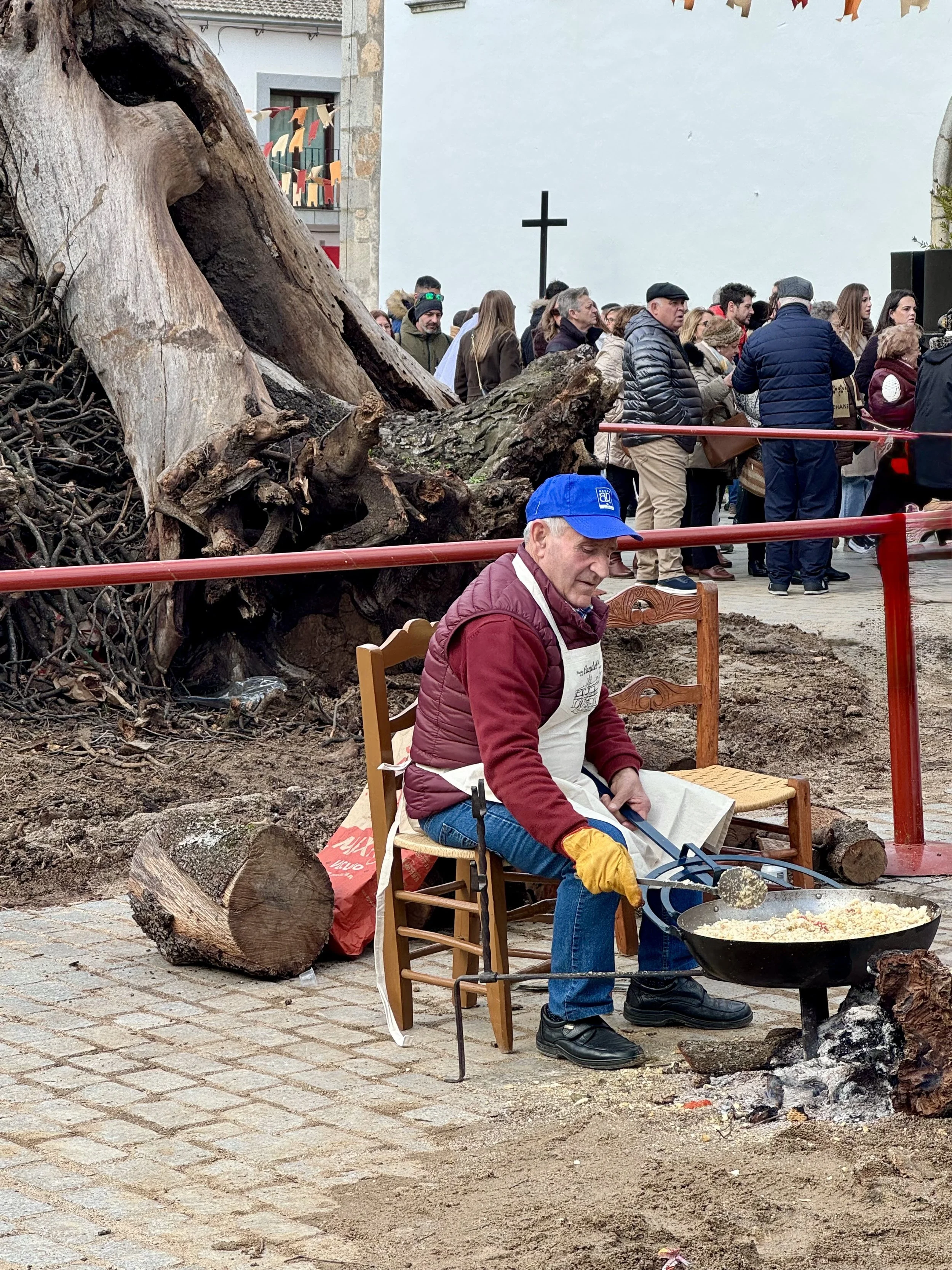 A man in a red jacket and blue cap cooking food in a black frying pan over a fire with ashes outside on a cobblestone street, with a large fallen tree and a group of people gathered in the background at an outdoor event.