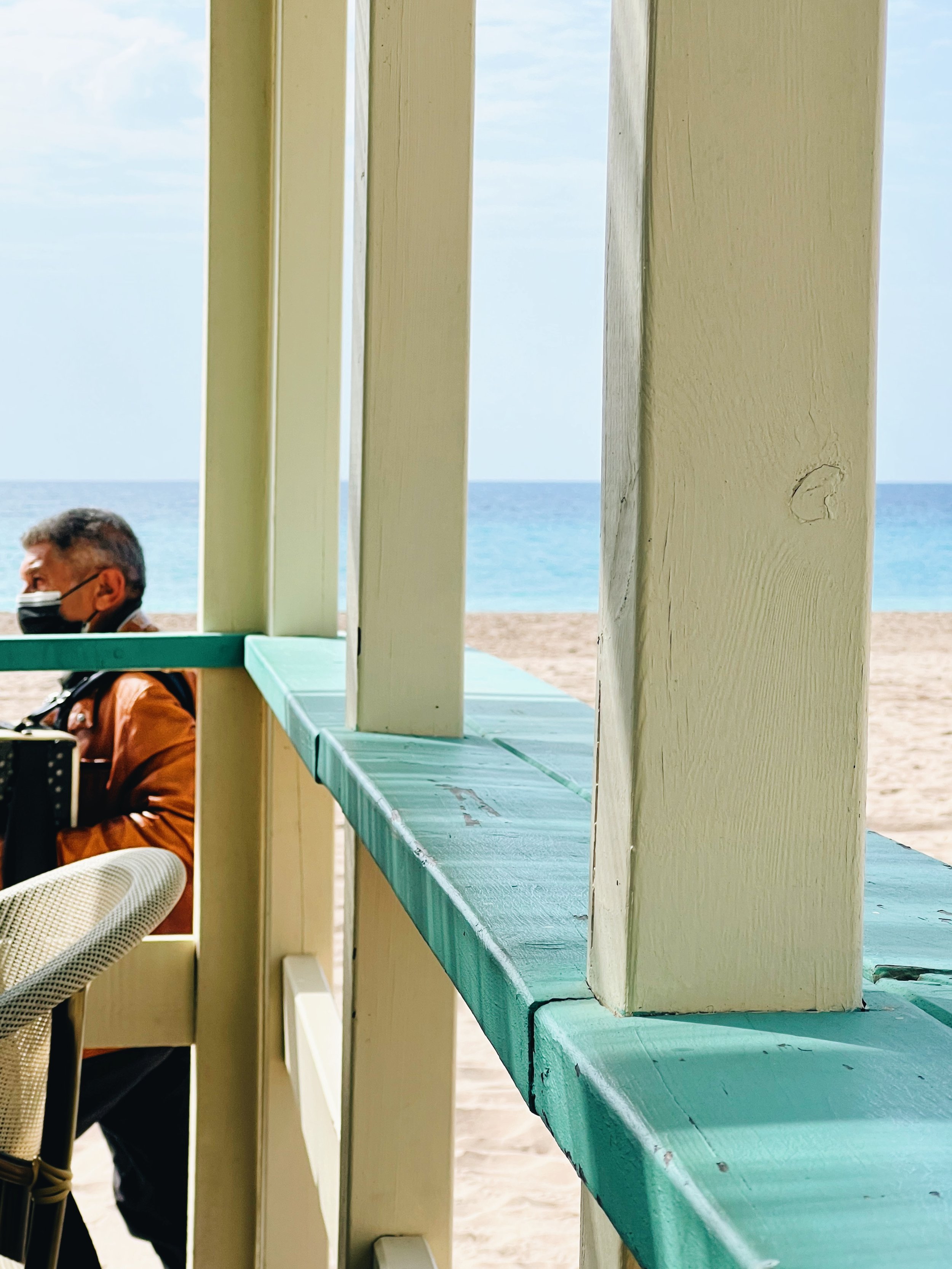 View of a beach scene through vertical wooden beams, with a man wearing a face mask sitting at a table in the lower left corner. The ocean and sky are visible in the background.