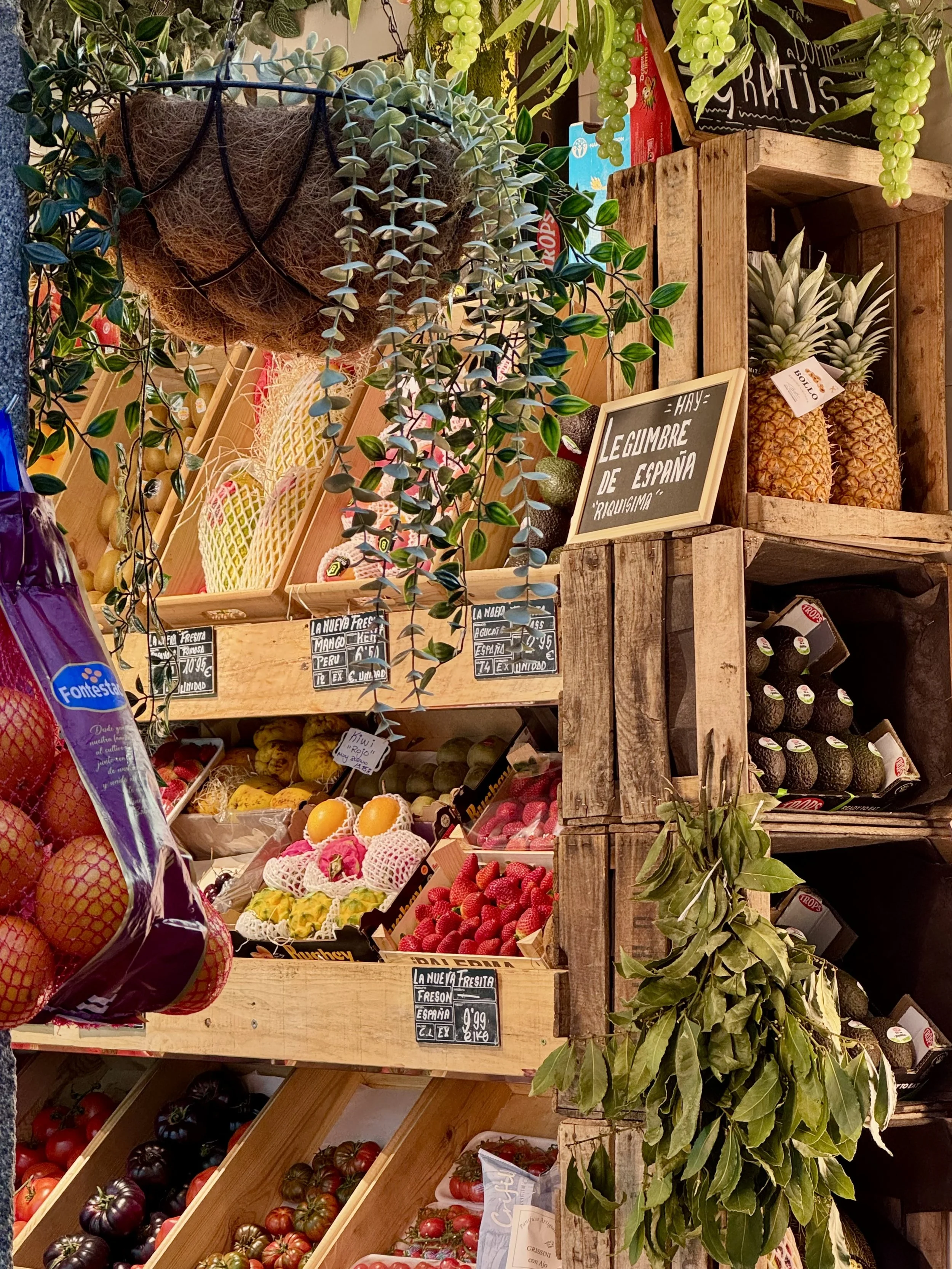 An assortment of fresh fruits including tomatoes, strawberries, pineapples, and melons displayed on wooden shelves at a market. Green leafy plants are also part of the display, along with handwritten signs in Spanish listing prices.