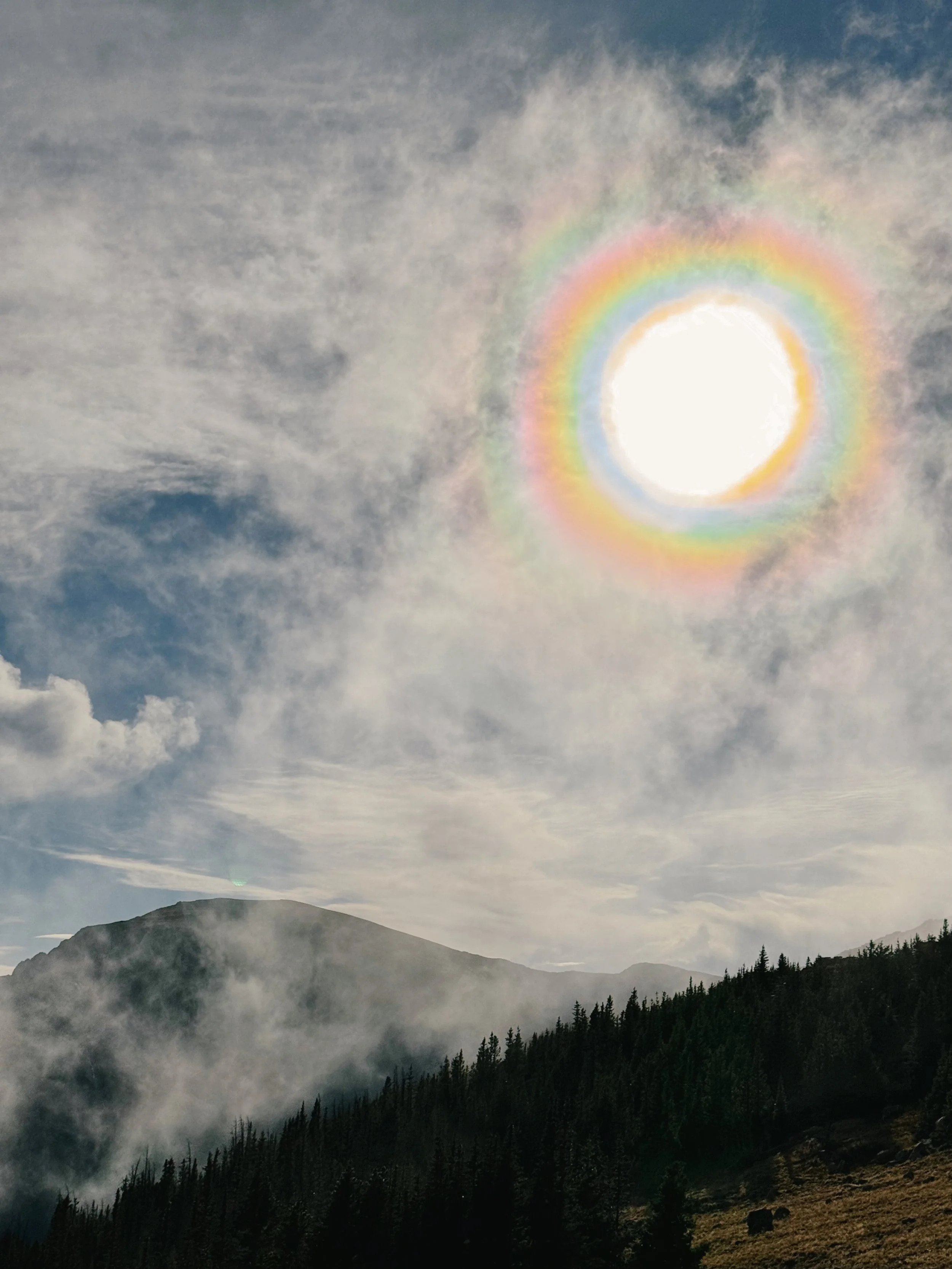 The sun with a rainbow halo appears behind clouds over a mountain landscape with pine trees and rocky terrain.