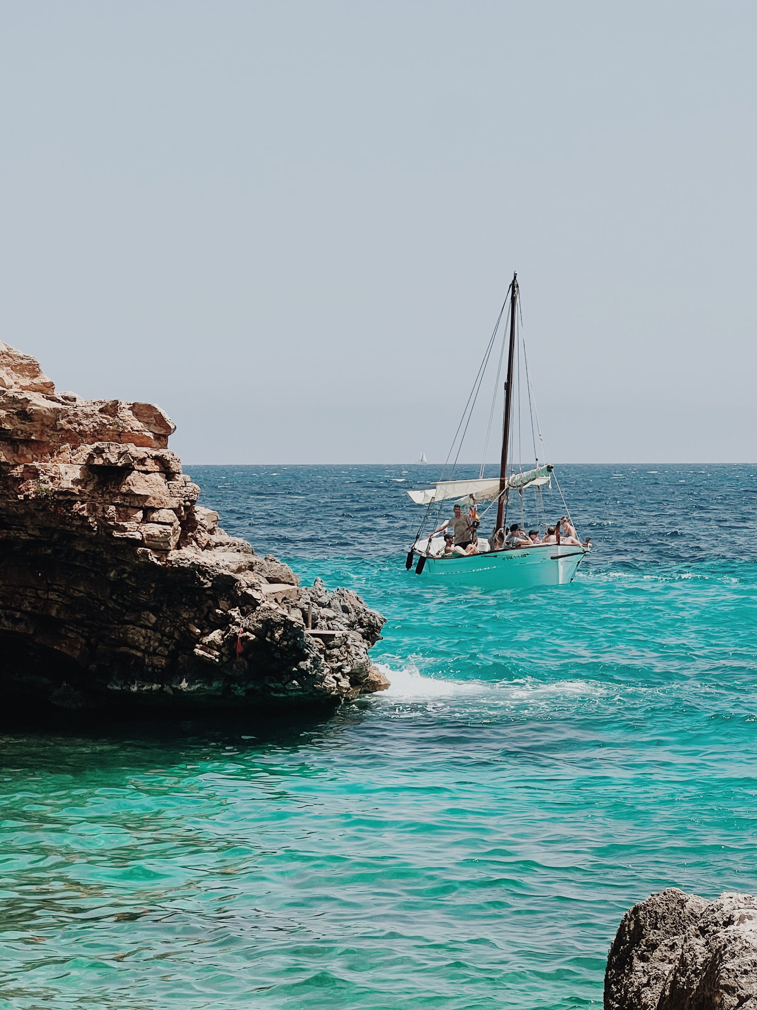 A boat with people onboard near a rocky coastline with clear blue-green water and a distant sailboat under a clear sky.
