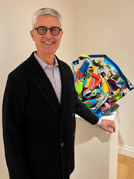 Doug Woodham standing next to a colorful abstract sculpture on a white pedestal in an art gallery.
