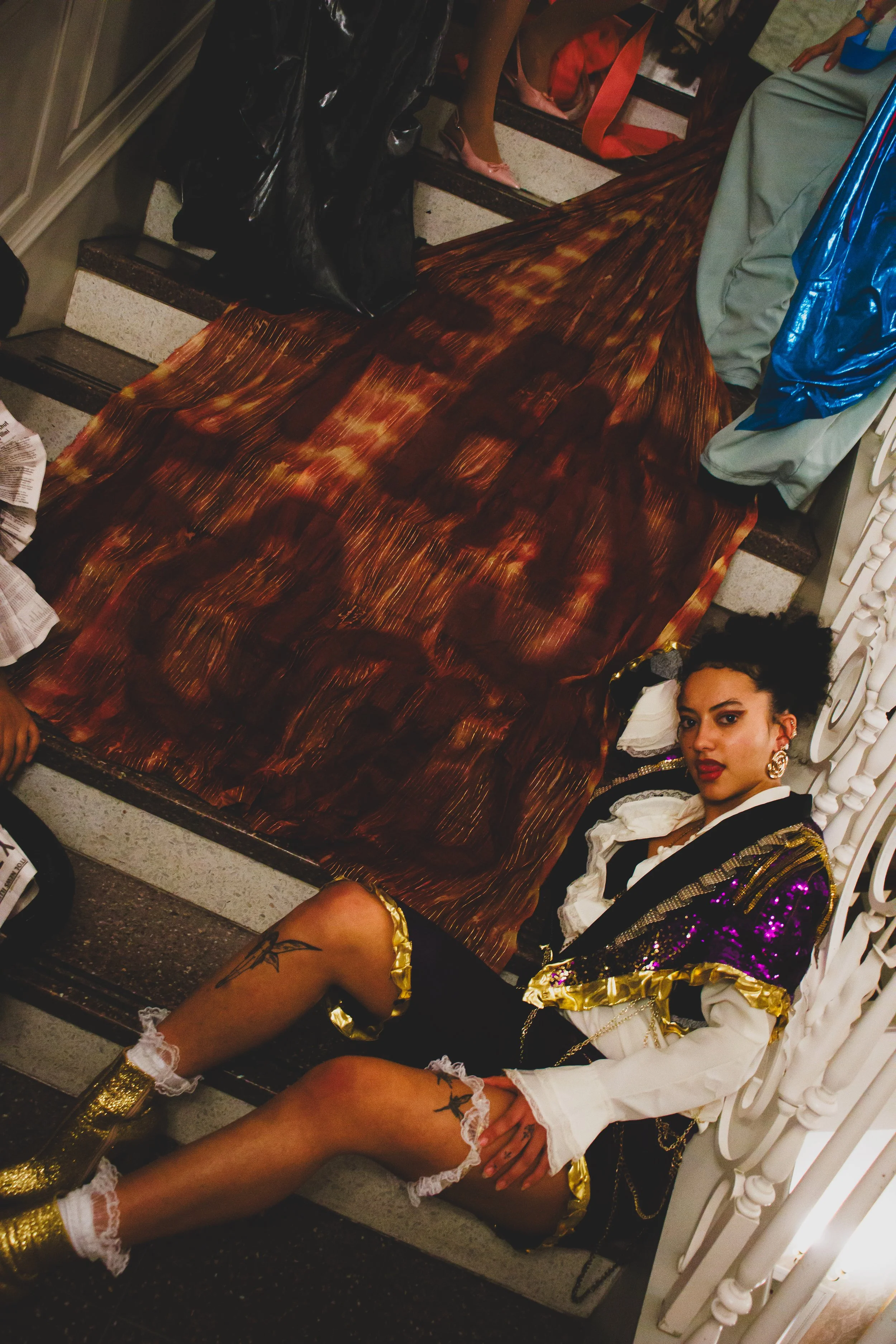 Woman in an elaborate black, white, gold, and purple costume reclining beside a large brown feathered prop indoors