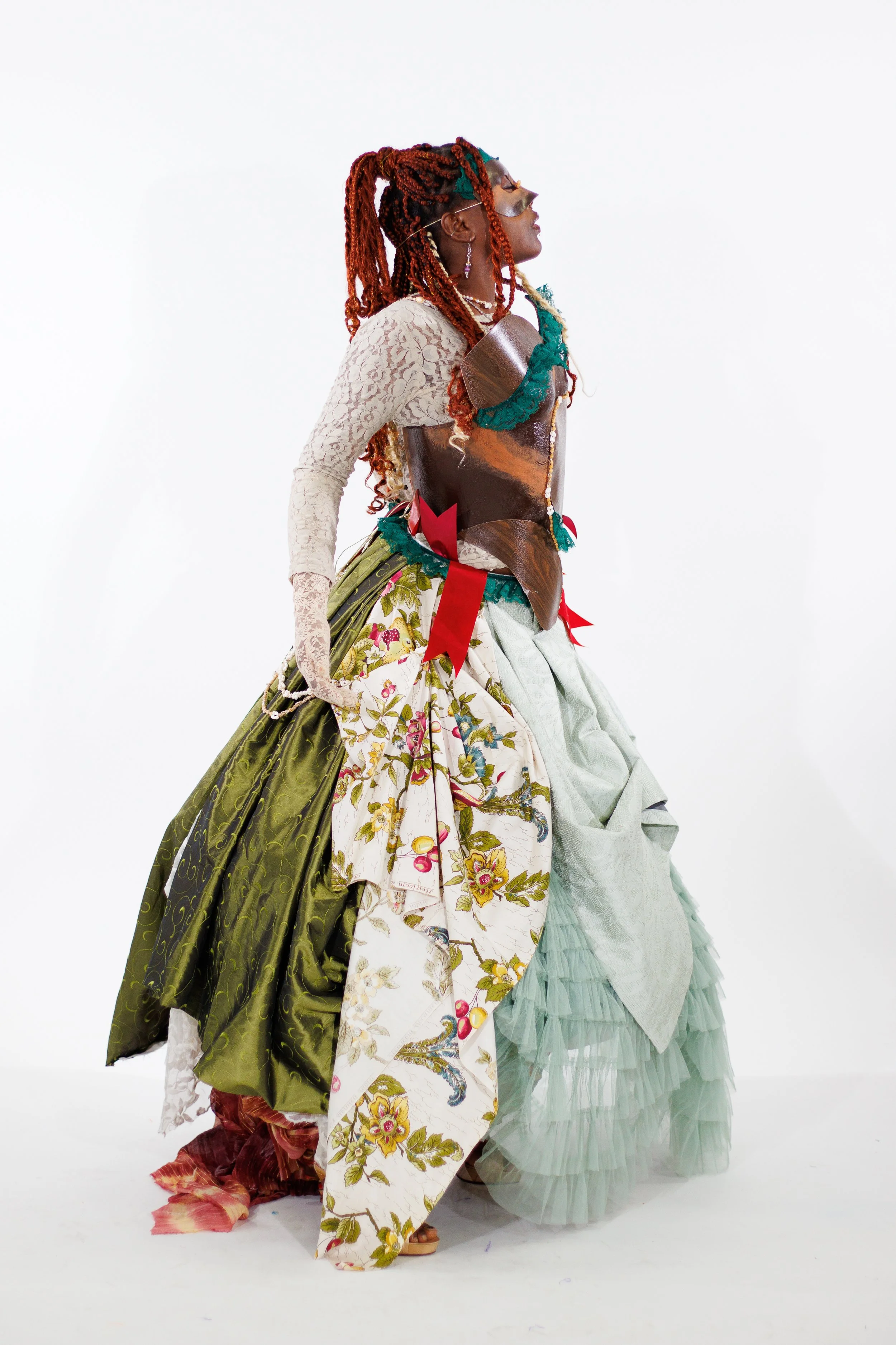 Person in profile wearing an elaborate theatrical costume with a metallic mask, red braids, floral skirt, and layered fabric drapery
