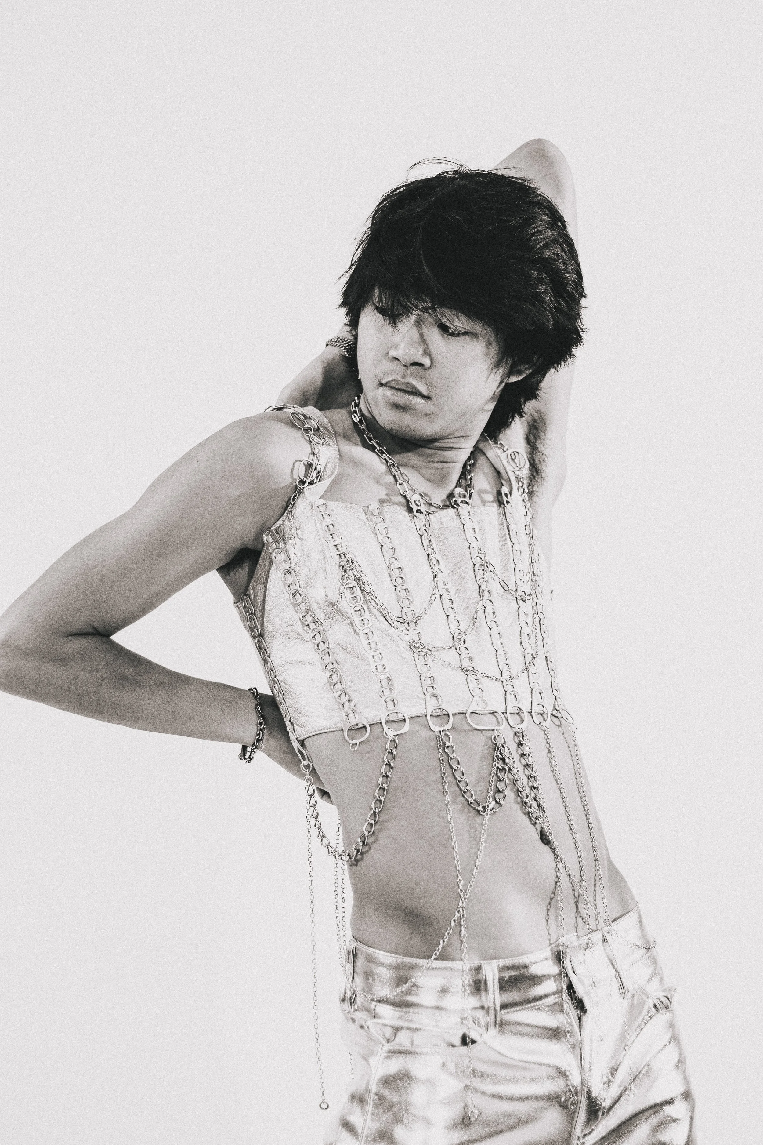 Black-and-white portrait of a person with short dark hair wearing layered chain jewelry and a metallic corset top.