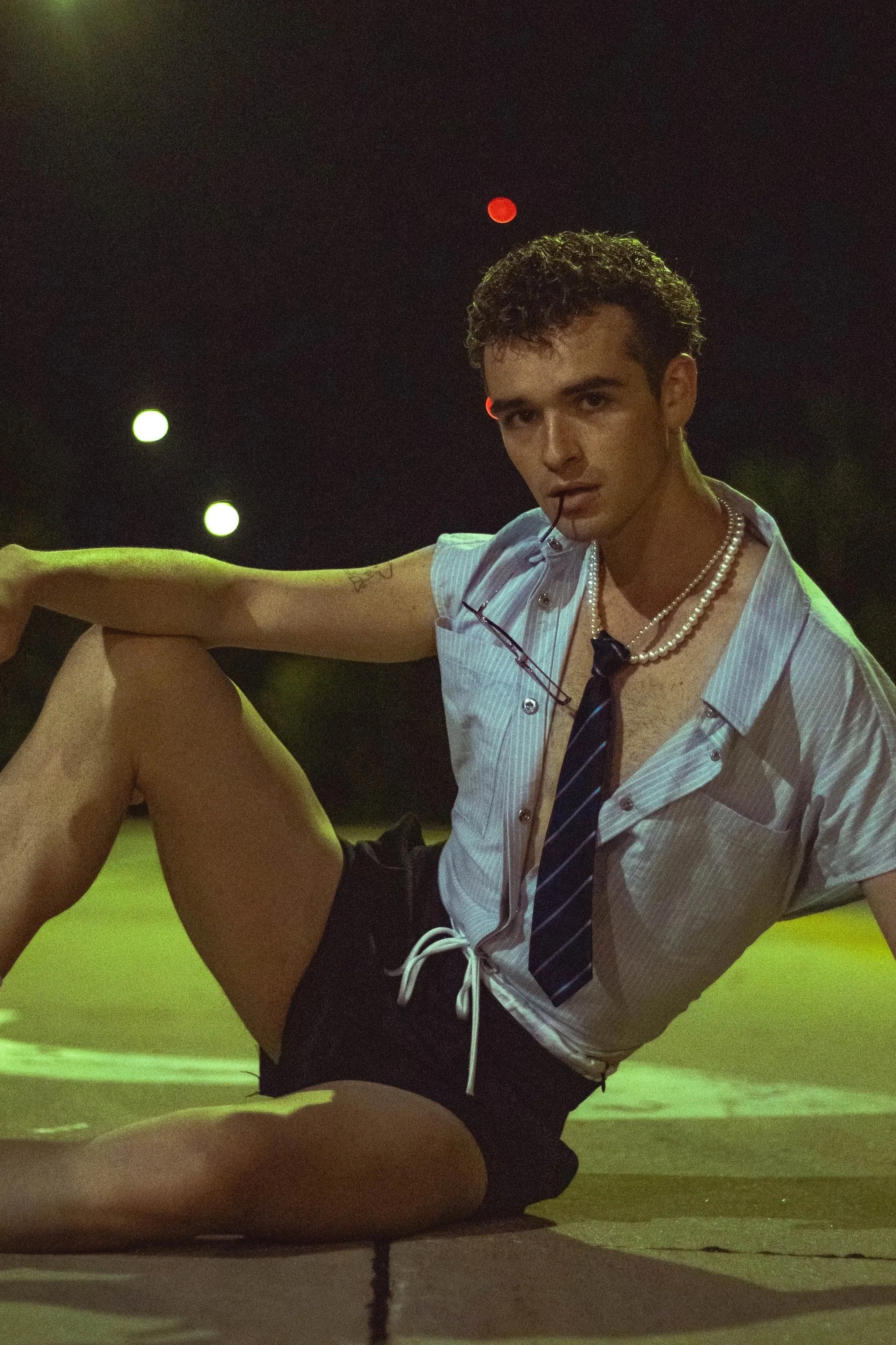 Man in a light blue shirt, striped tie, and pearl necklace kneeling on a dimly lit stage.