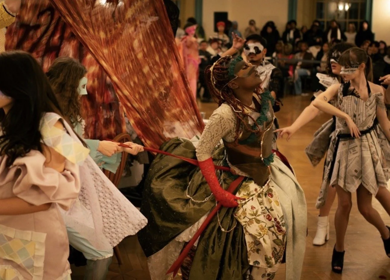 Masked performers in elaborate costumes dance at a lively indoor masquerade ball.
