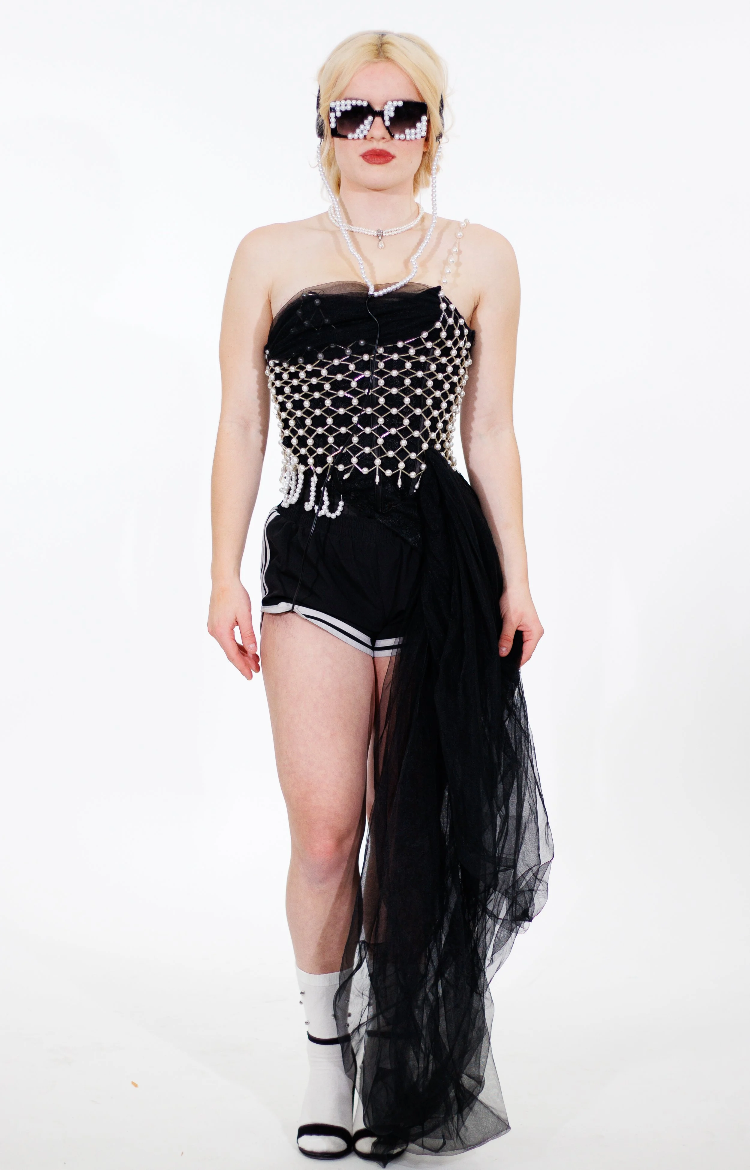 Fashion model wearing pearl-embellished sunglasses, layered pearl jewelry, and a black sheer tulle dress against a white background