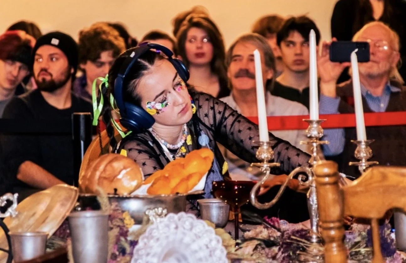 Costumed diner wearing headphones and face paint at a candlelit Halloween feast, surrounded by spectators.