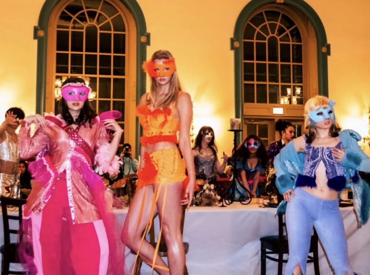 Masked fashion models in colorful costumes pose at a masquerade party in an ornate dining hall.