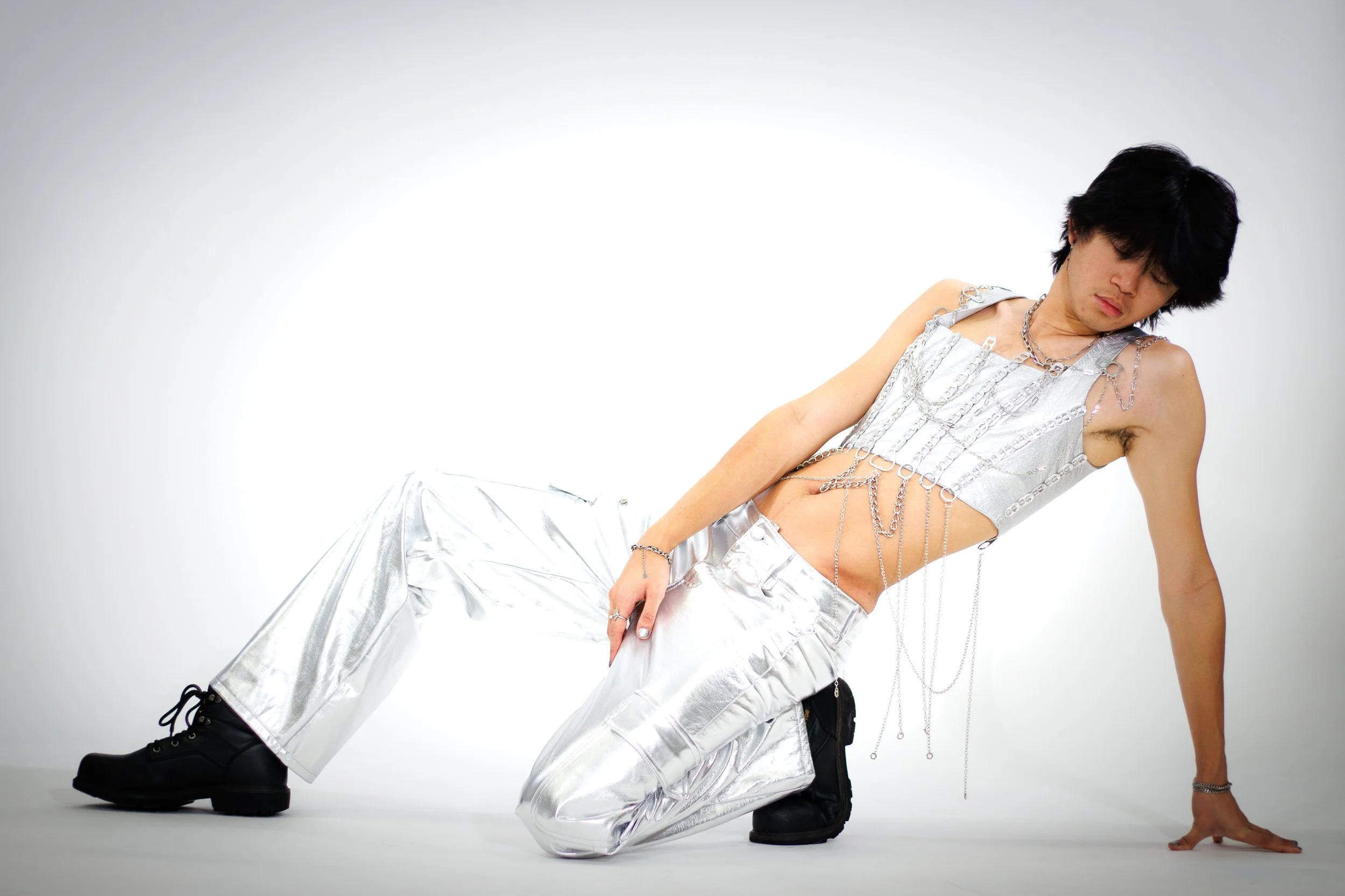 Fashion model posing in a silver chain-adorned crop top, metallic pants, and black platform boots against a white studio background