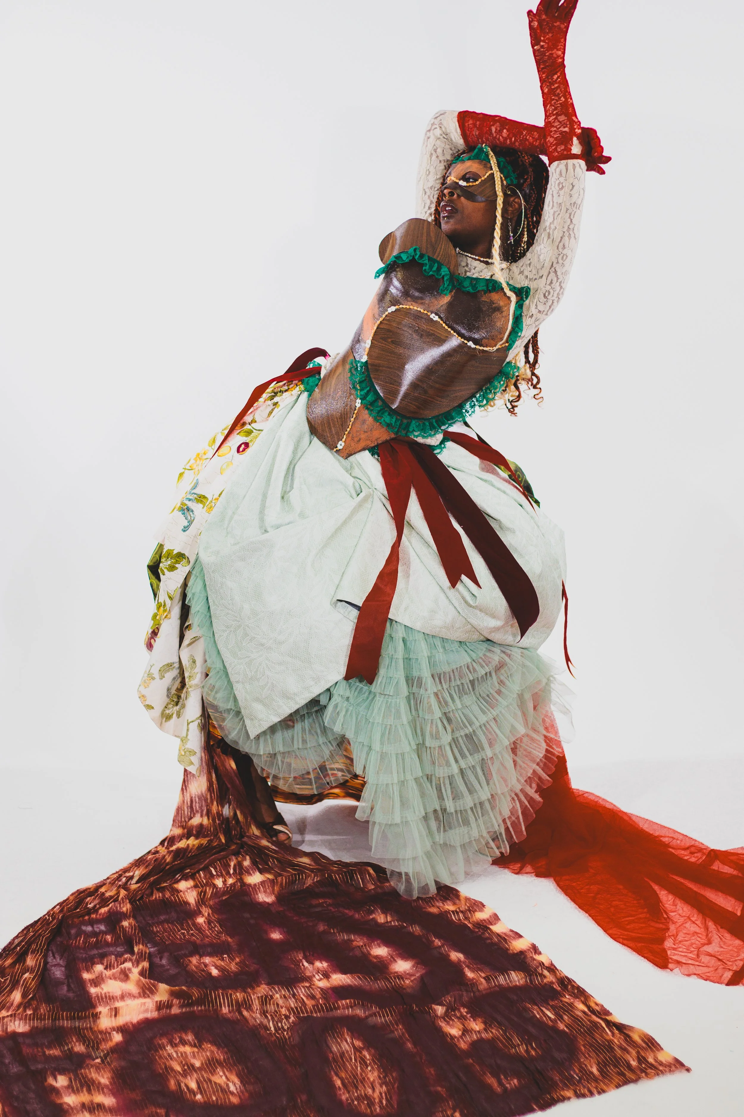 Black performer in an elaborate colorful costume, mask, and lace sleeves posing with one arm raised against a white background