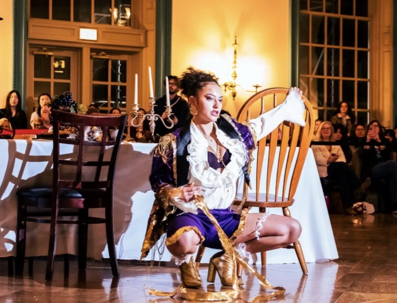 Theatrical performer in an ornate costume kneeling beside a wooden chair during a dinner theater performance