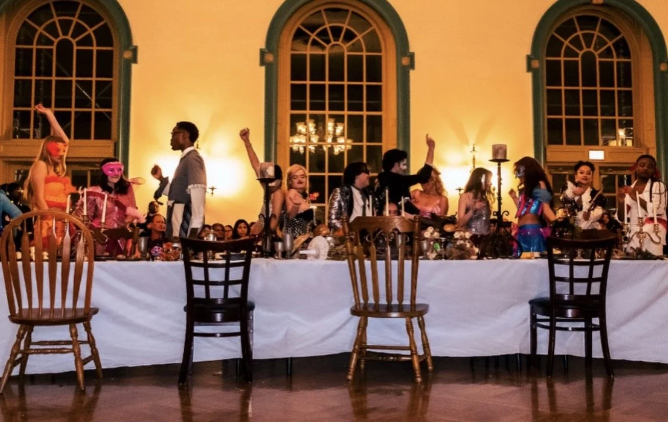 Costumed guests wearing masks celebrate at a formal masquerade party around a long banquet table.