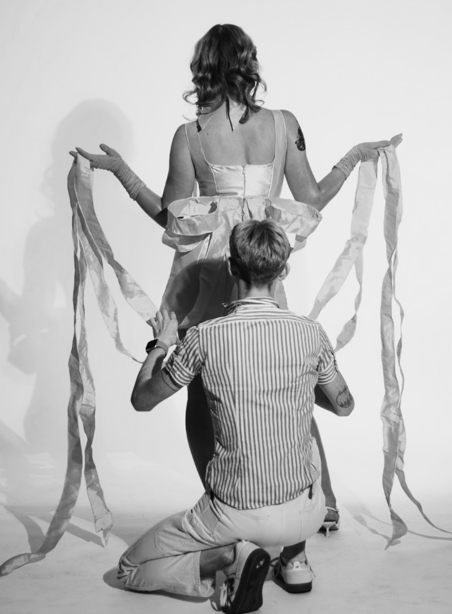 Black-and-white photo of a person kneeling behind a woman in a dress holding long flowing ribbons.