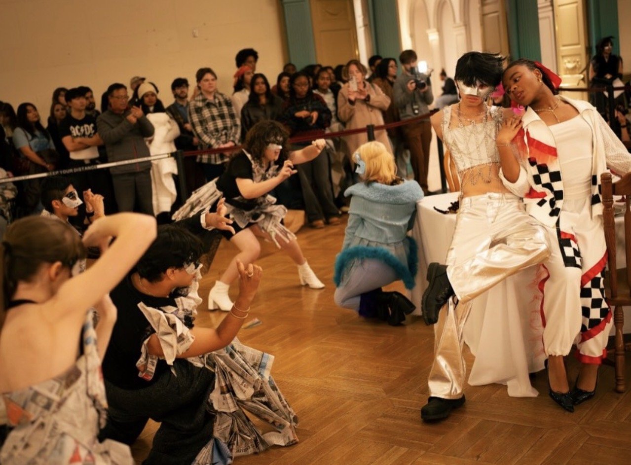 Costumed performers pose and dance before a crowd at an indoor fashion or art event.
