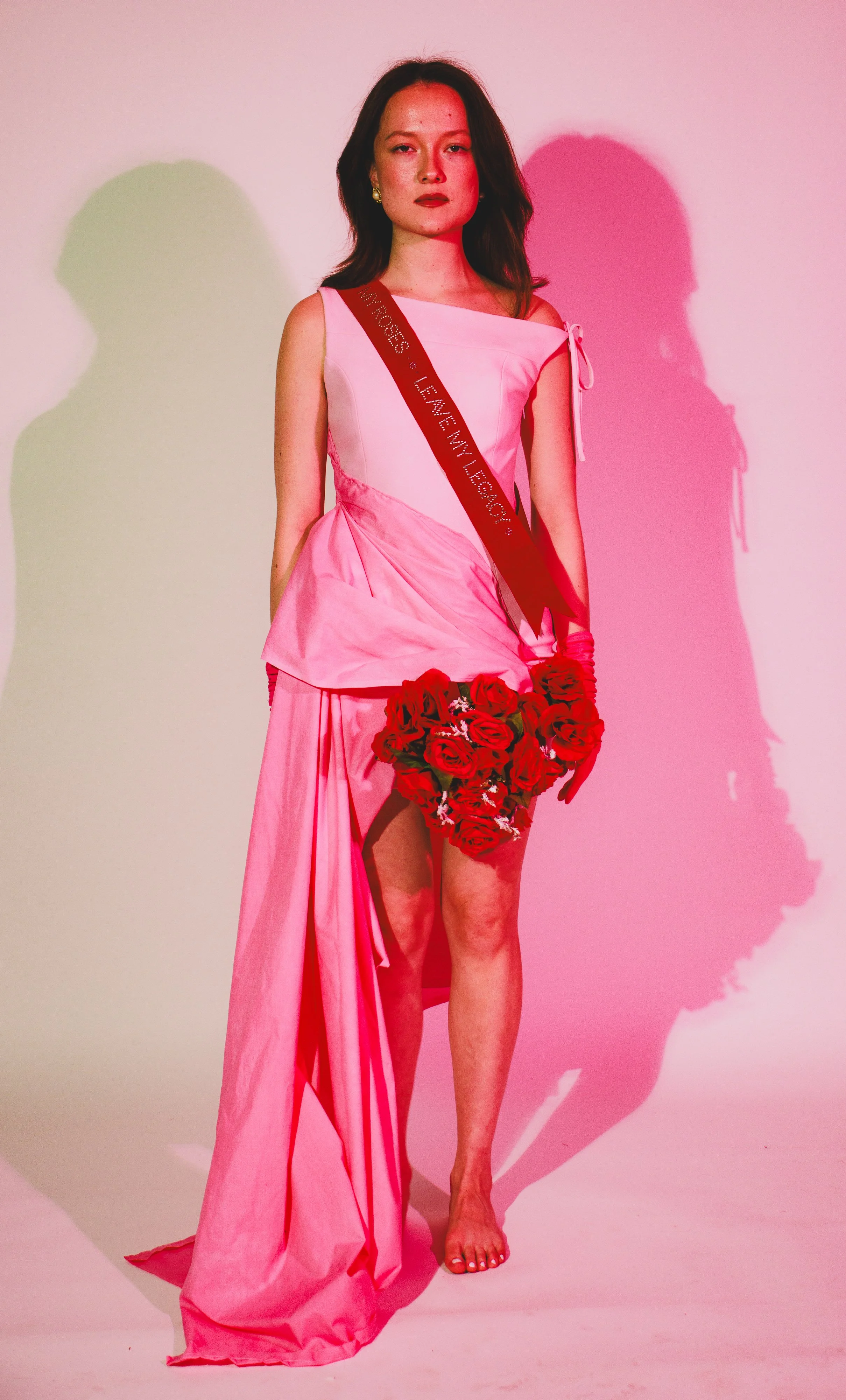 Woman in a pink dress wearing a red sash and holding a bouquet of red roses against a pink background