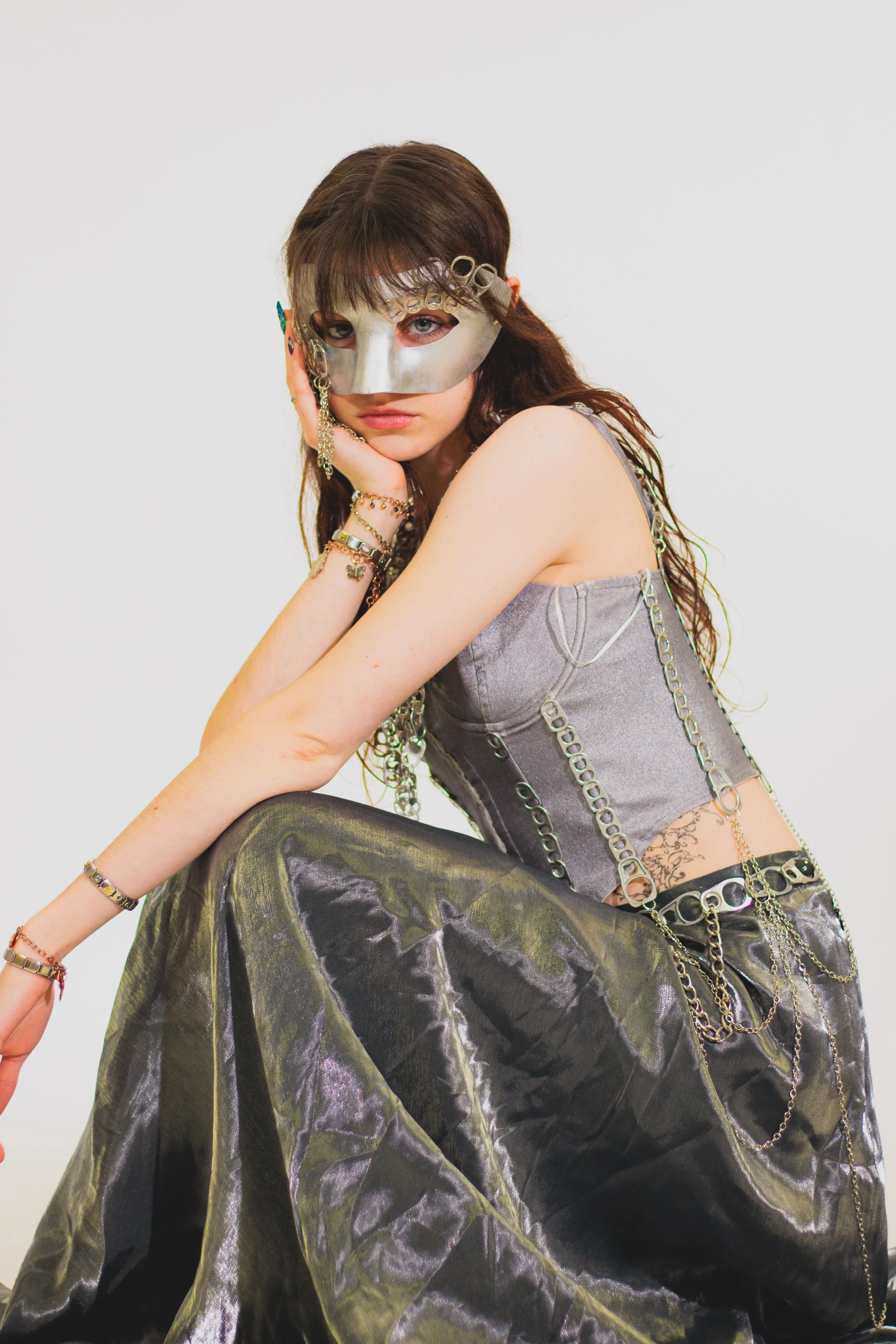 Person with long brown hair wearing an ornate metallic mask, gray corset, layered chains, and black satin pants
