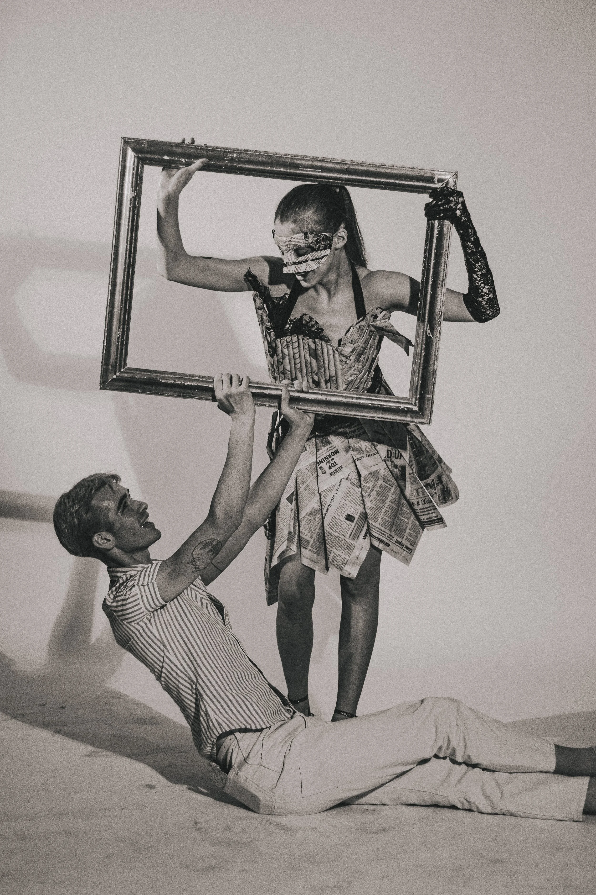 Black-and-white theatrical scene with a woman in a newspaper dress and lace gloves holding a picture frame while a man lies on the floor beneath her.