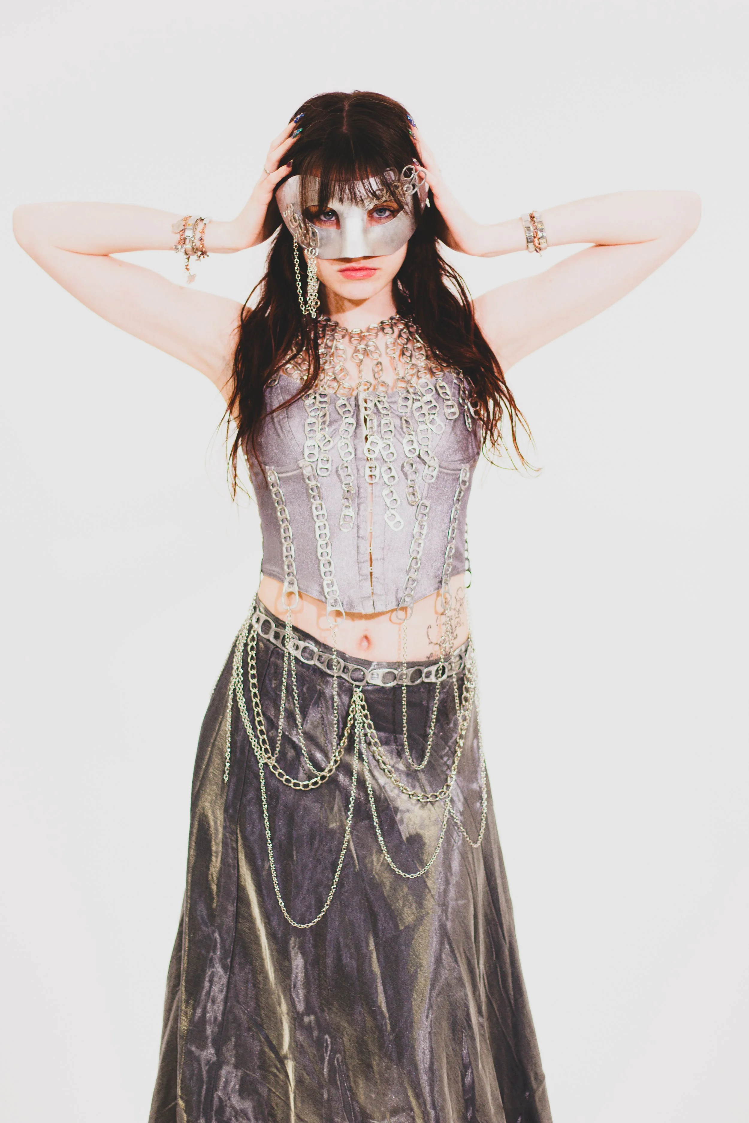 Fashion portrait of a woman with metallic face paint, dark hair, and layered chain embellishments posing against a white background