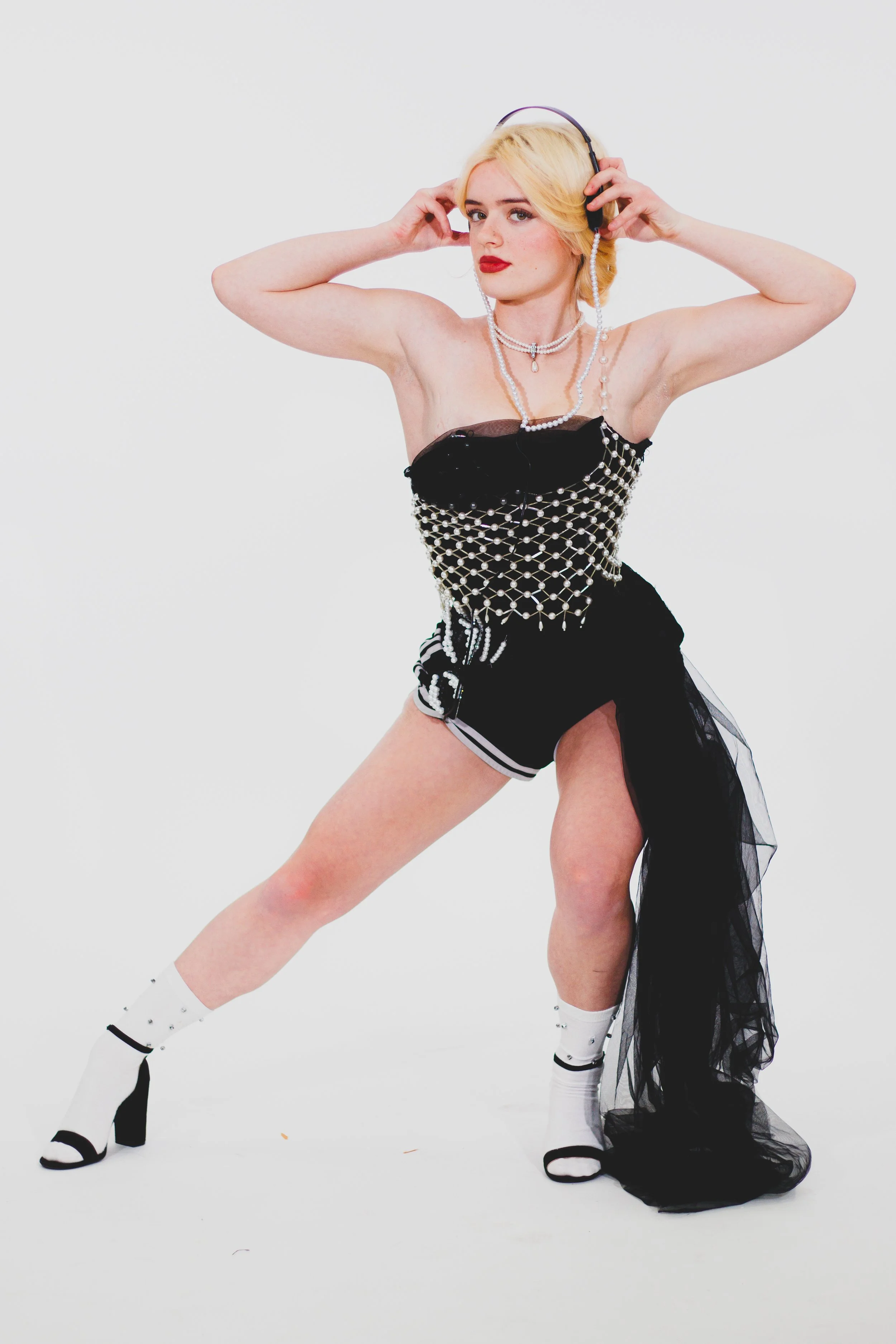 Blonde woman in a pearl-studded black outfit adjusting headphones against a white background